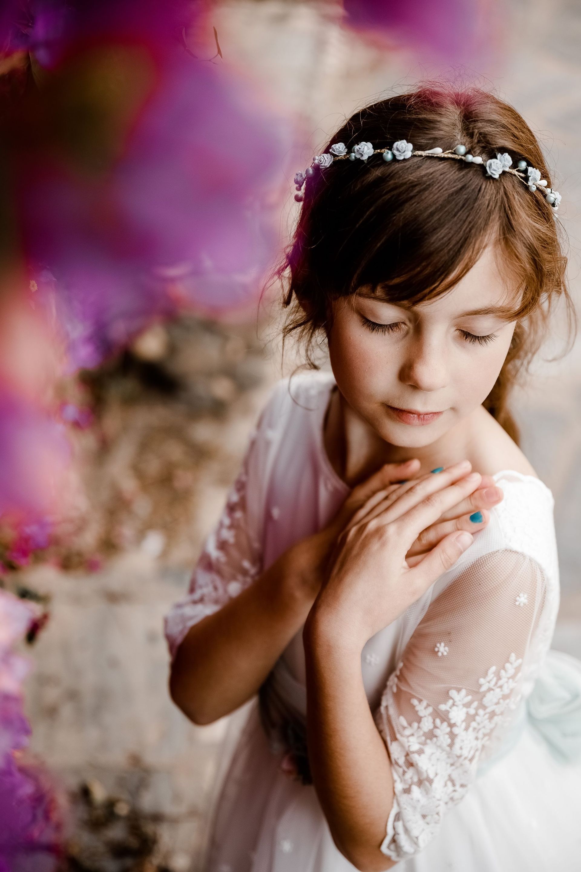 Niña pequeña con un vestido blanco de encaje mirando hacia abajo, con las manos sobre los hombros y una diadema floral sobre un fondo rosa pálido.