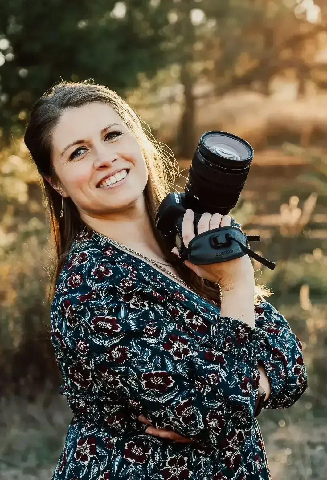 Mujer sonriente al aire libre, sosteniendo una cámara réflex digital al hombro bajo la cálida luz del sol.