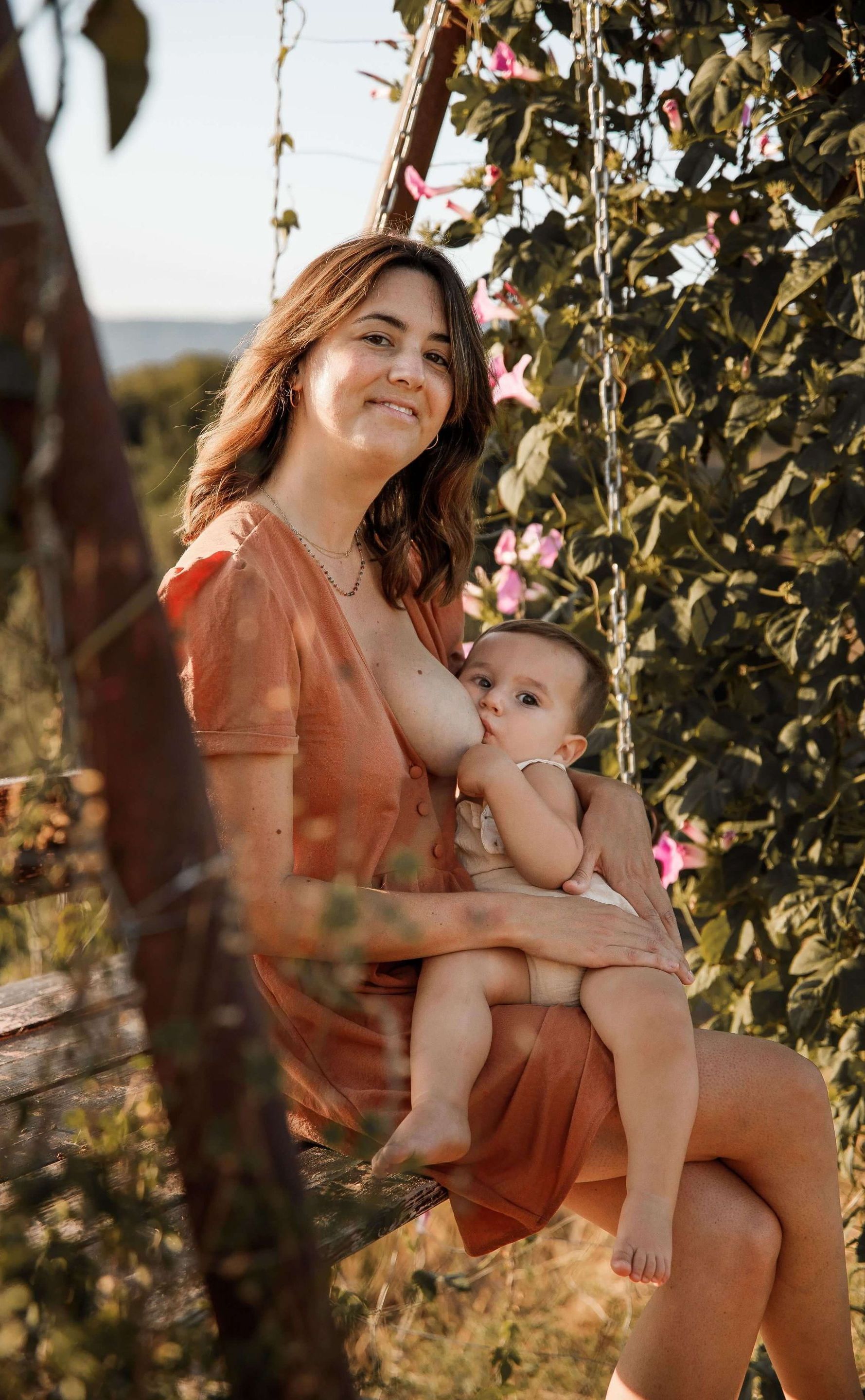 Mujer con vestido naranja sosteniendo a un bebé en un columpio hecho con un árbol, al aire libre, rodeada de vegetación.