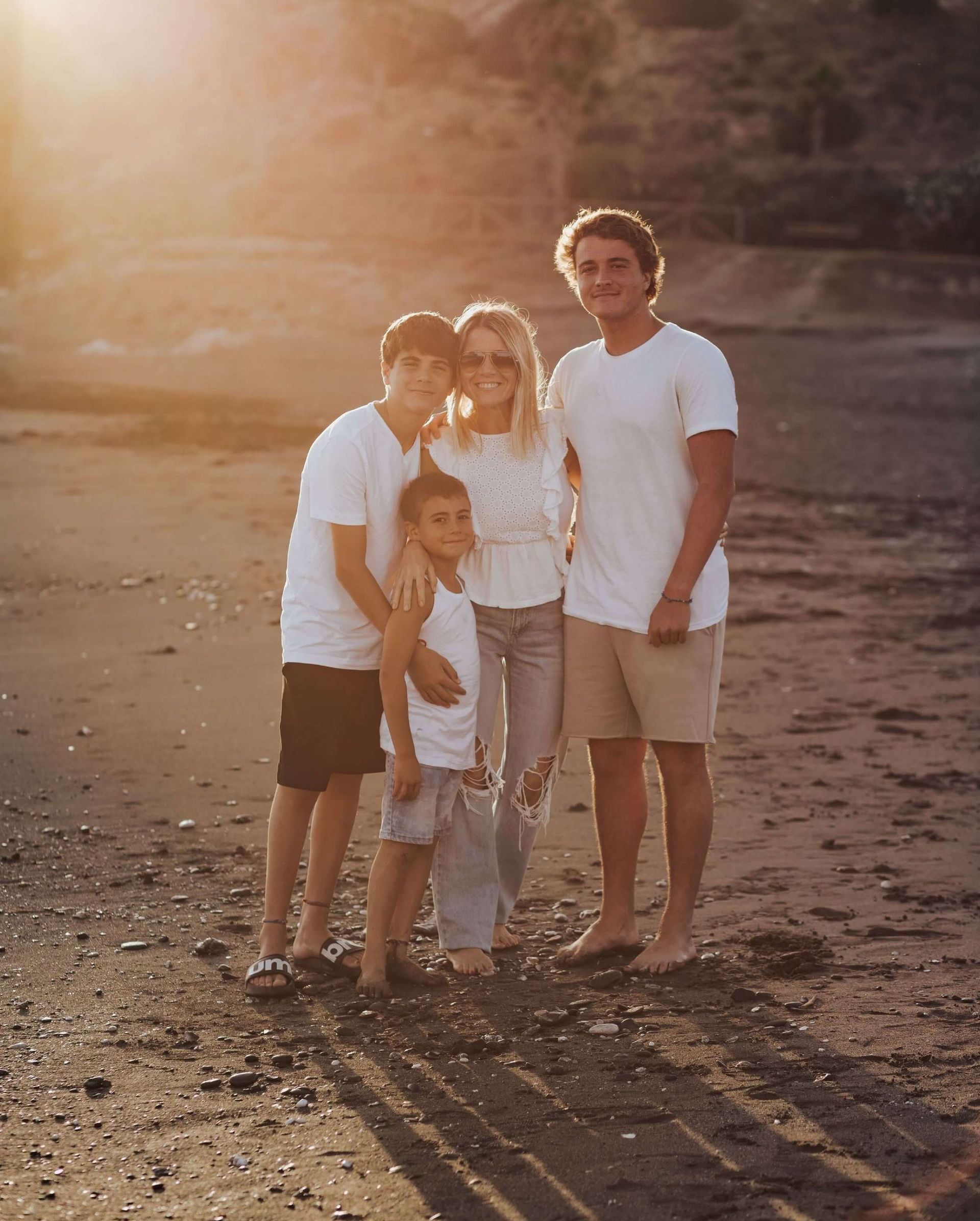 Cuatro personas vestidas de blanco de pie juntas en una playa al atardecer.