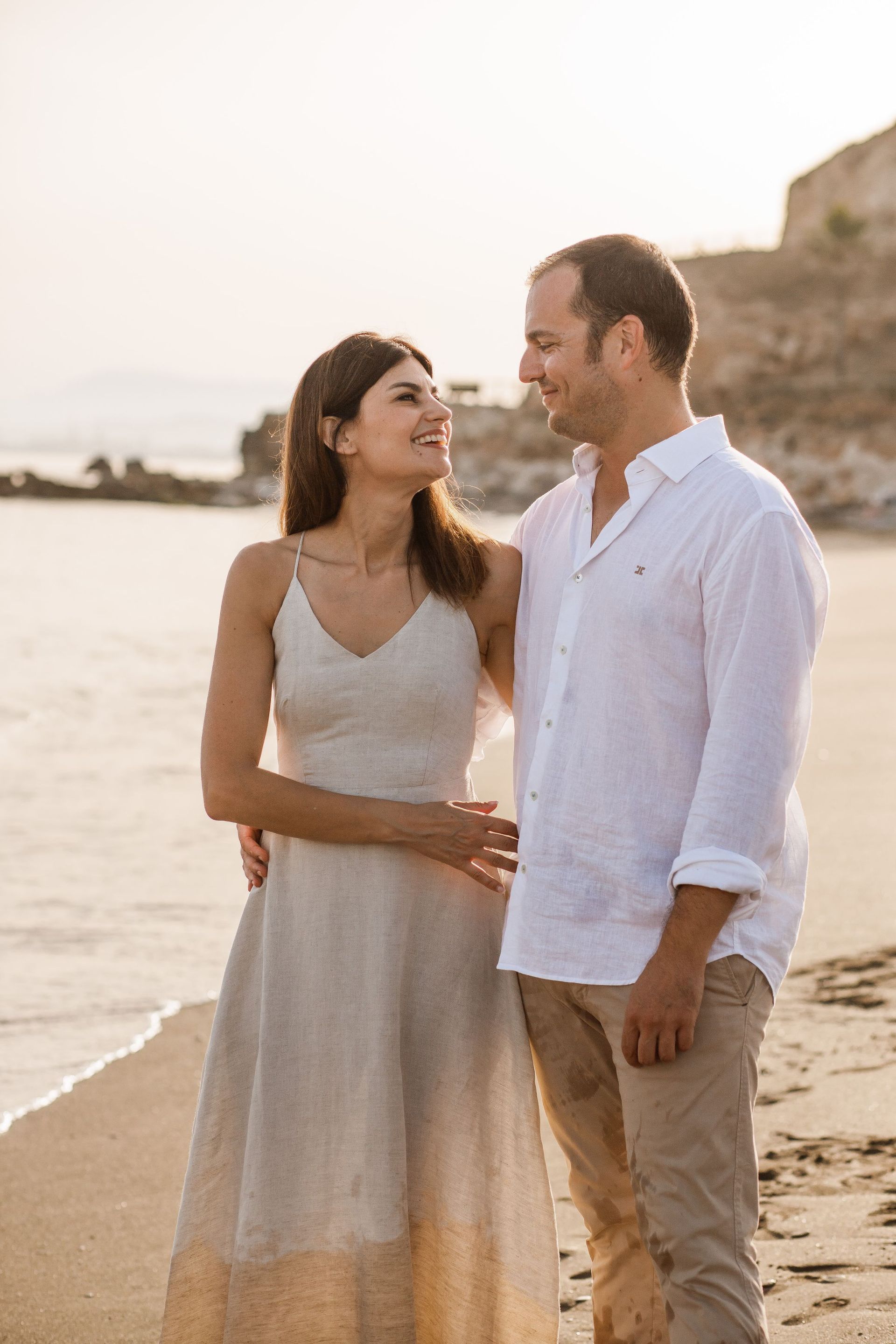 Una pareja vestida con ropa clara está de pie en una playa al atardecer, sonriéndose mutuamente.