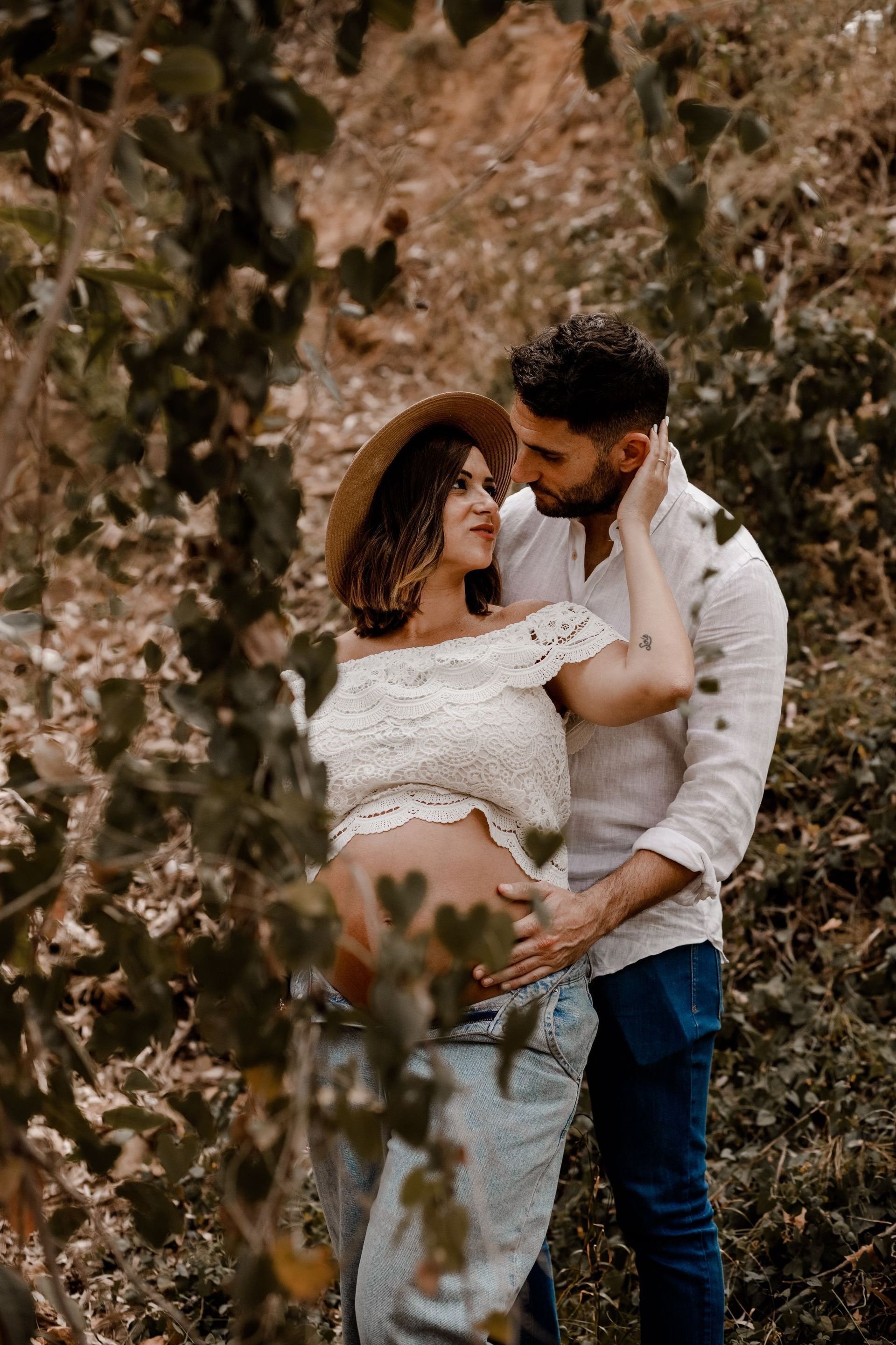 Una pareja de pie en una zona boscosa, abrazados y mirándose el uno al otro, con el suave follaje otoñal de fondo.