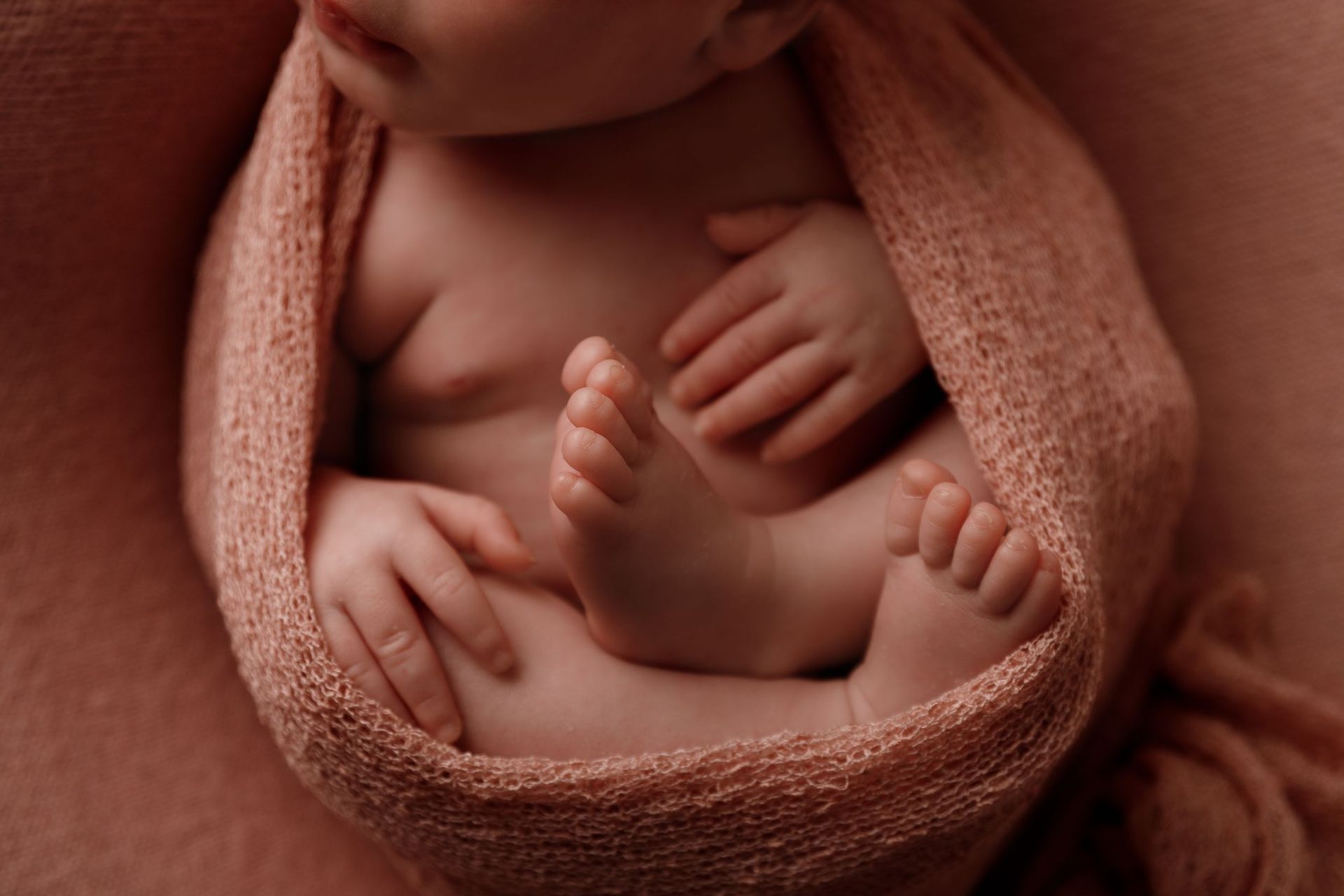 Bebé recién nacido envuelto en una manta tejida de color rosa, con manos y pies visibles.