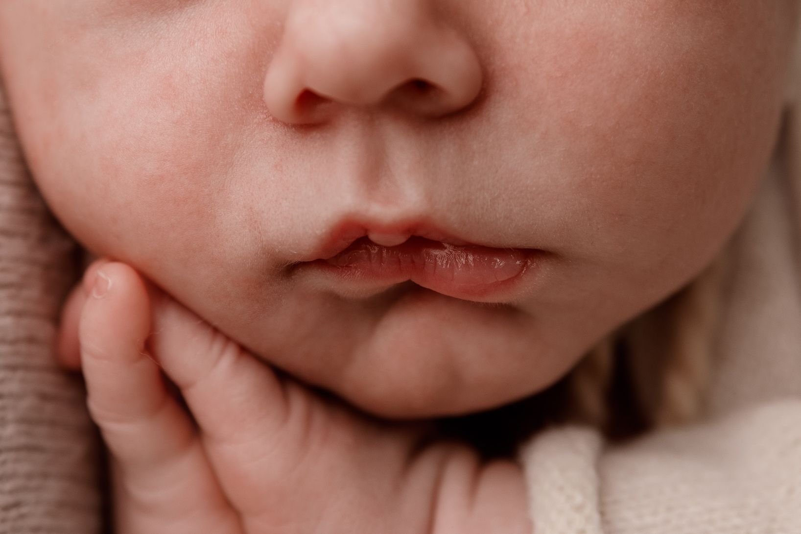 Primer plano del rostro de un bebé dormido con una mano cerca de la boca, envuelto en una suave tela color crema.
