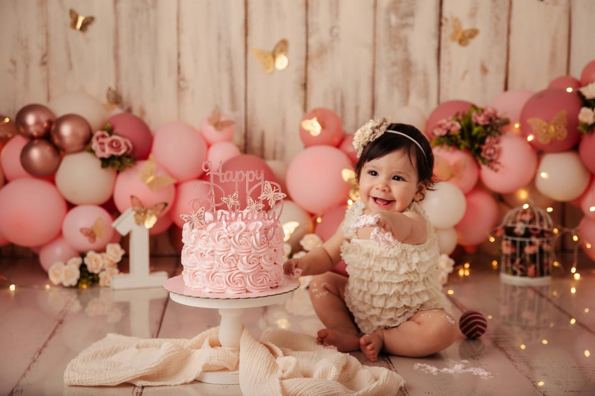 Un bebé sentado junto a un pastel de cumpleaños rosa en una escena de fiesta decorada con globos de color oro rosa.