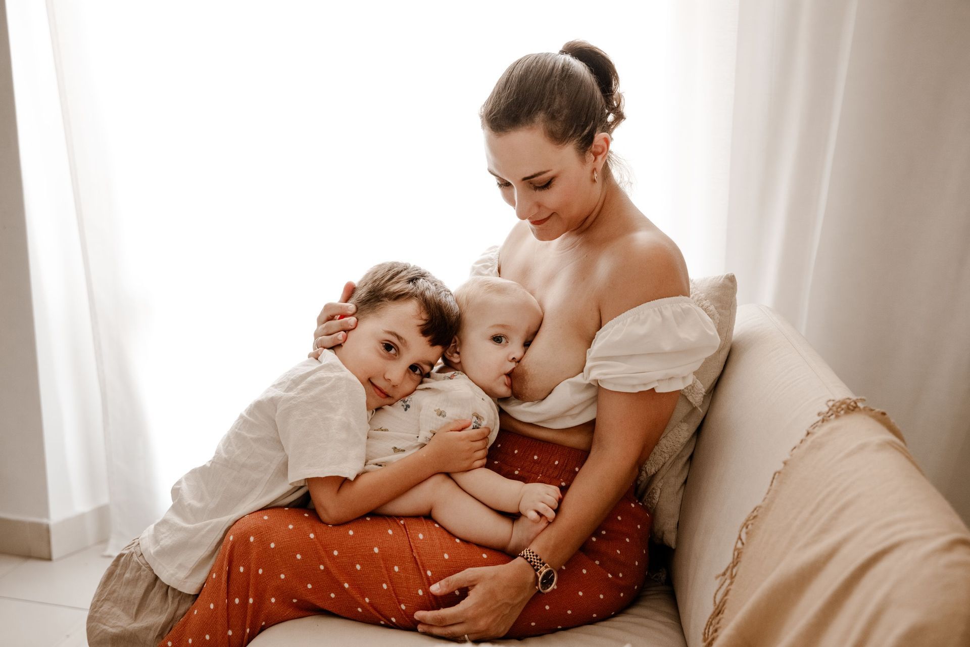 Mujer amamantando en un sofá beige con dos niños, abrazándolos en una habitación luminosa.
