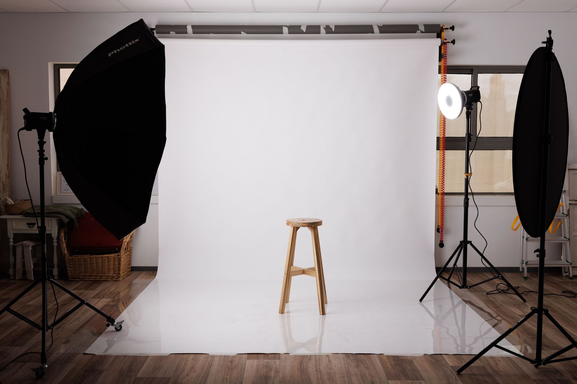 Montaje de estudio fotográfico con fondo blanco, dos focos softbox y un taburete de madera en el centro.
