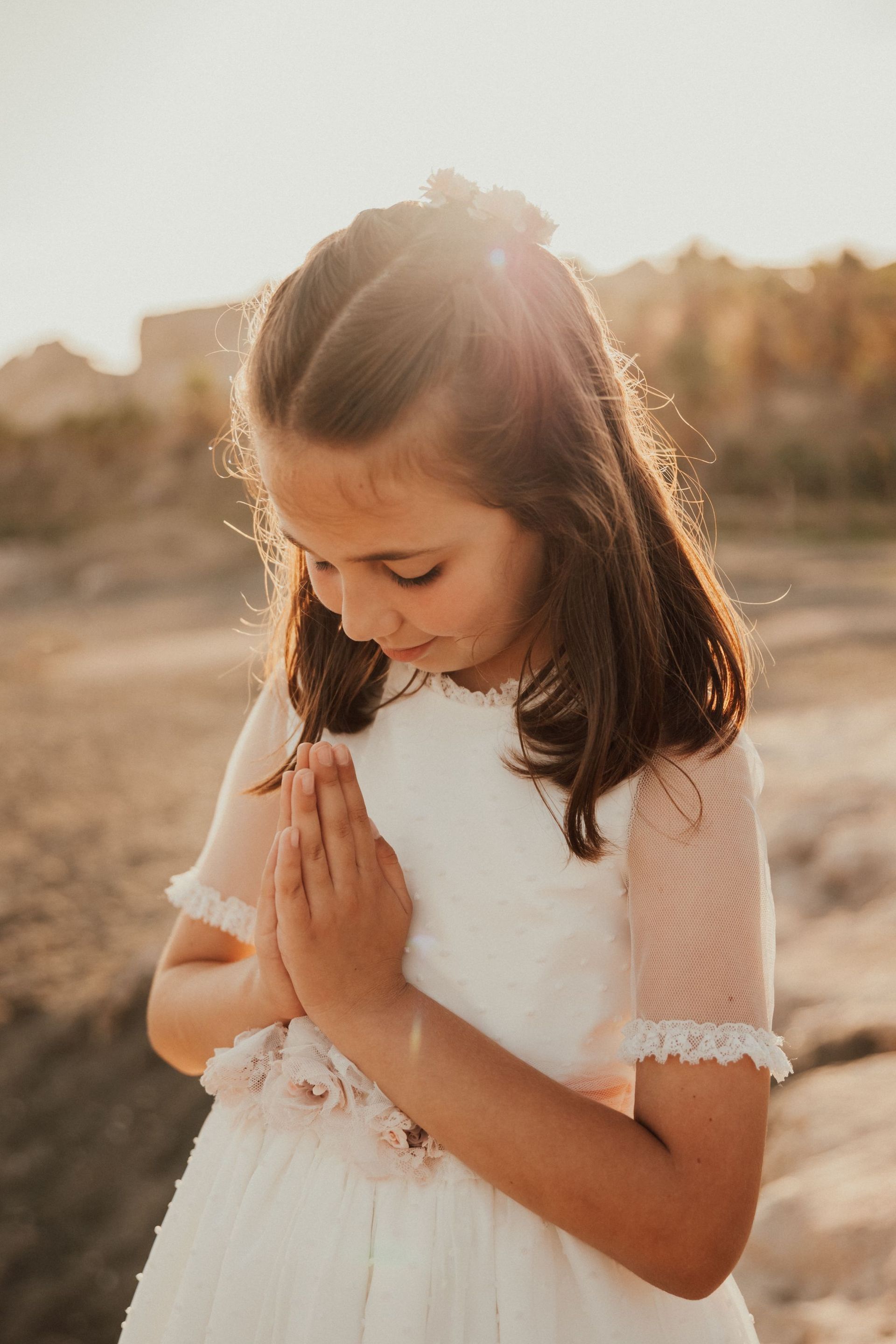 Niña vestida de blanco con las manos juntas en señal de oración, de pie al aire libre bajo la cálida luz del sol.