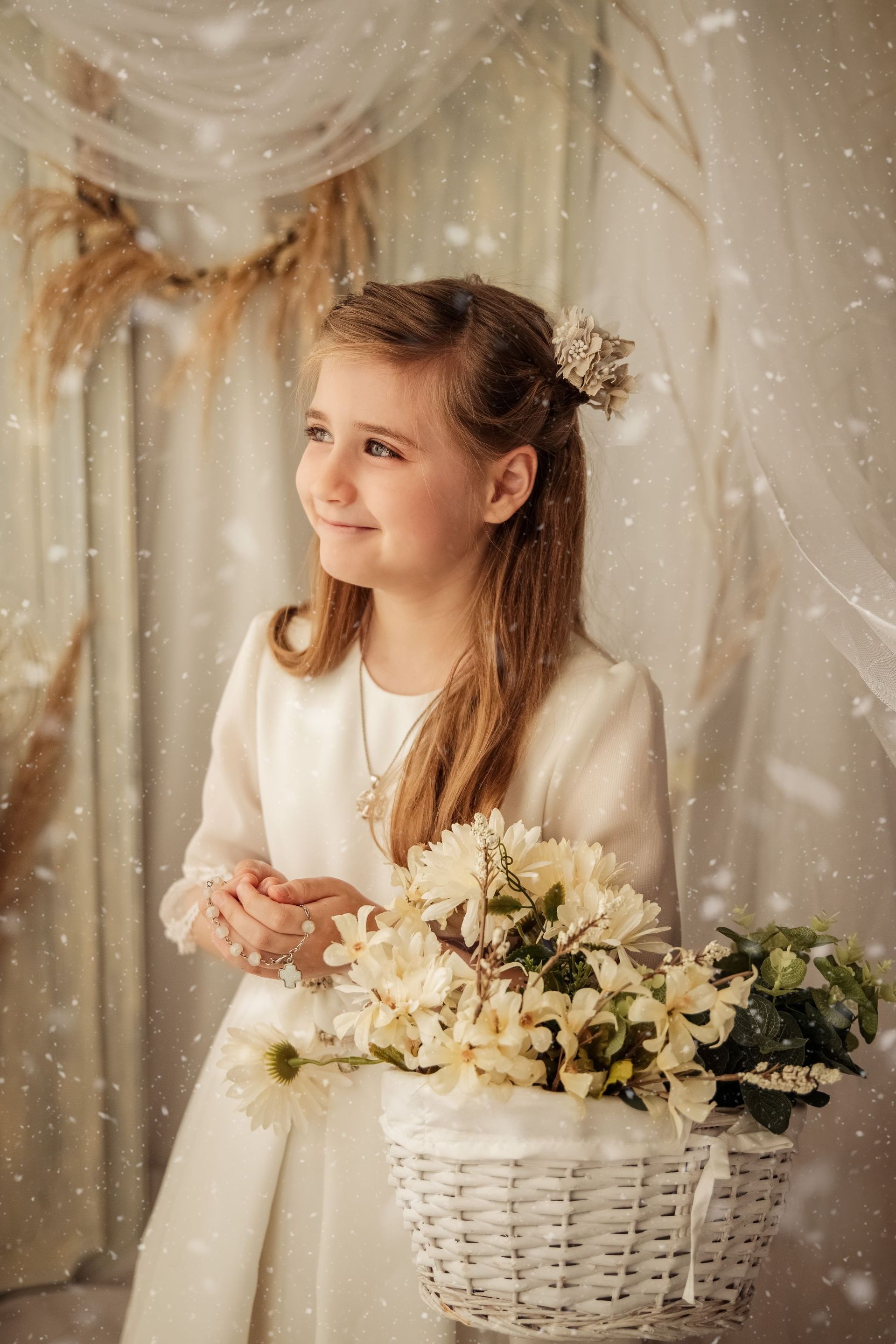 Una joven vestida de blanco, junto a una cesta de mimbre llena de flores, sonríe en un ambiente apacible y festivo.