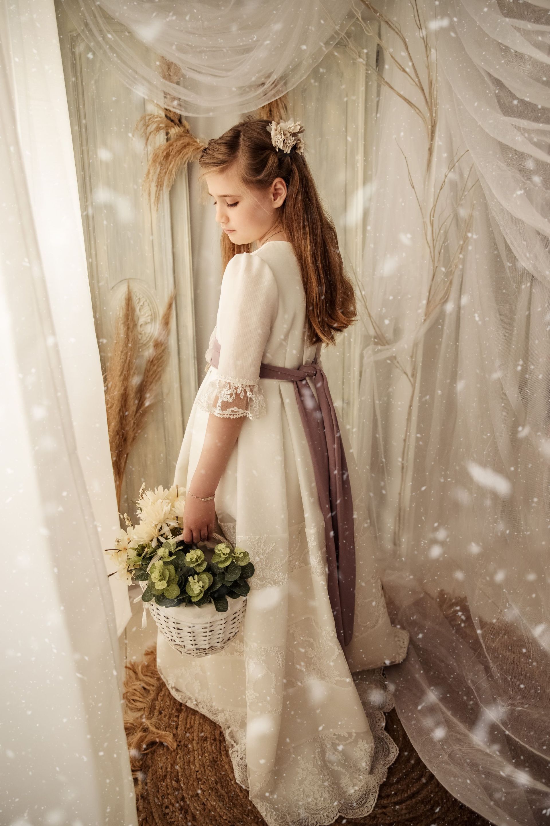 Una joven con un vestido blanco sostiene un ramo de flores junto a unas cortinas transparentes en una habitación con iluminación suave y cálida.