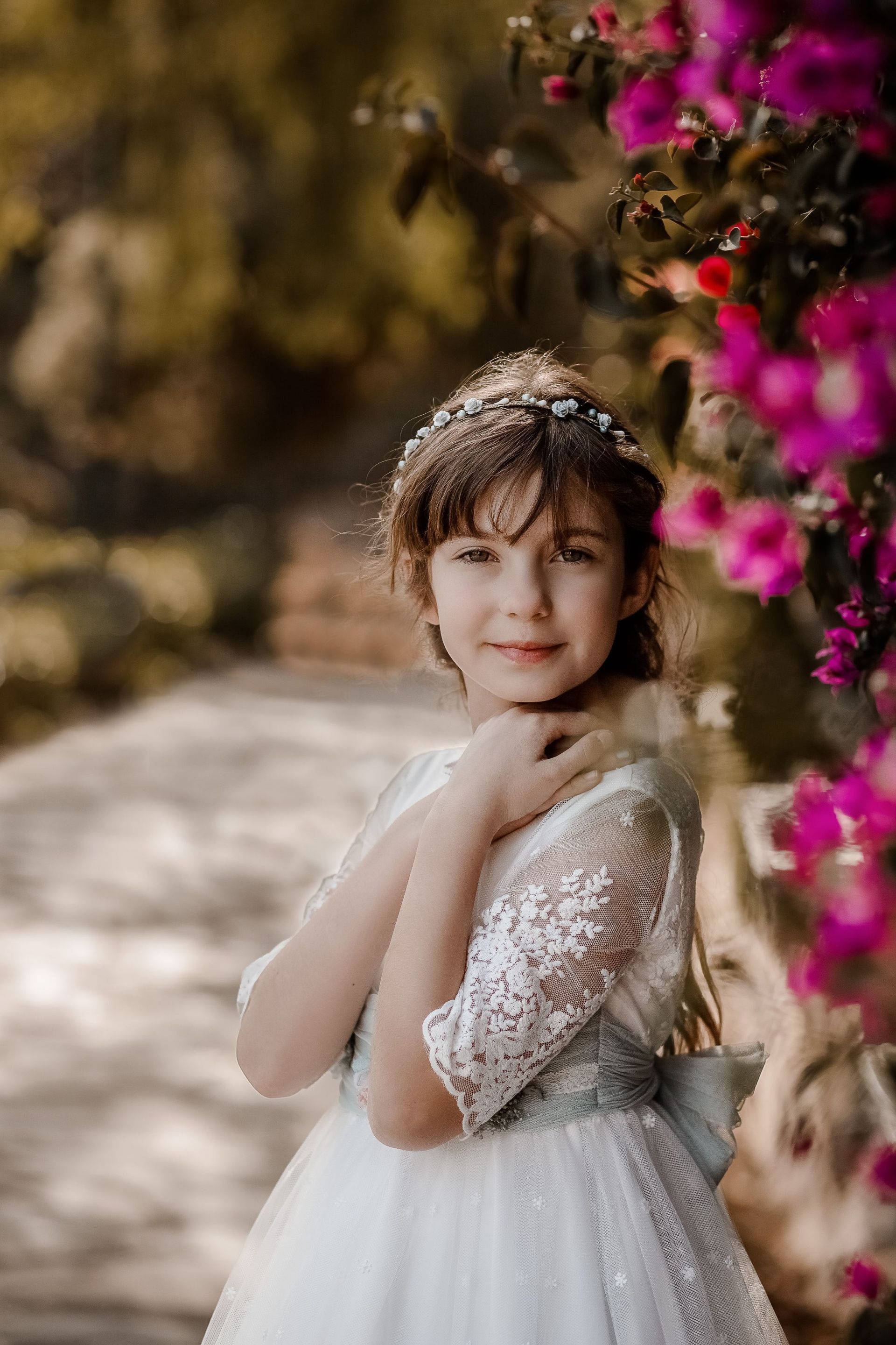 Niña con vestido blanco y tocado floral, de pie junto a flores rosas al aire libre.