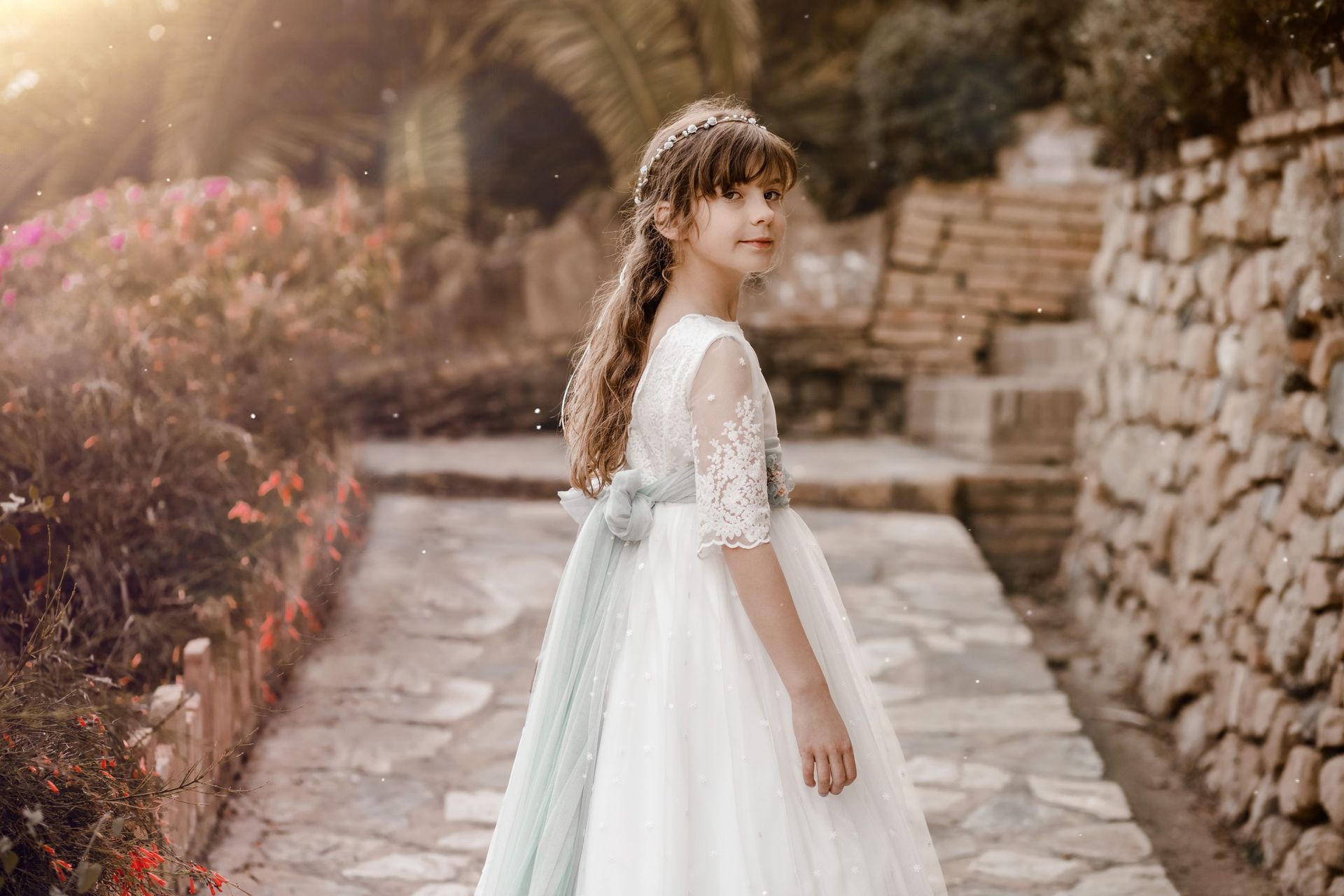 Una niña con un vestido blanco está de pie en un sendero de piedra junto a un muro de ladrillos en un entorno ajardinado.