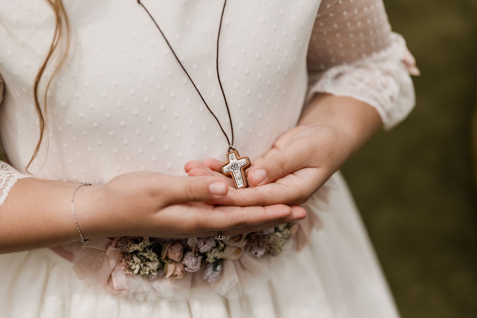 Manos que sostienen un pequeño colgante sobre un vestido blanco con mangas de encaje y un cinturón floral.