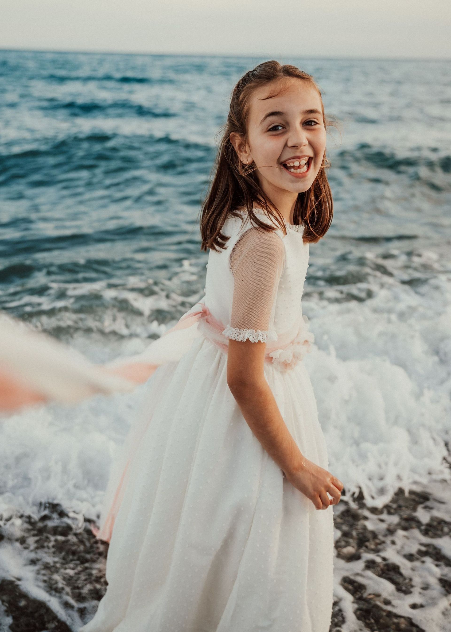 Niña sonriente con un vestido blanco de pie en una playa rocosa con olas detrás de ella.