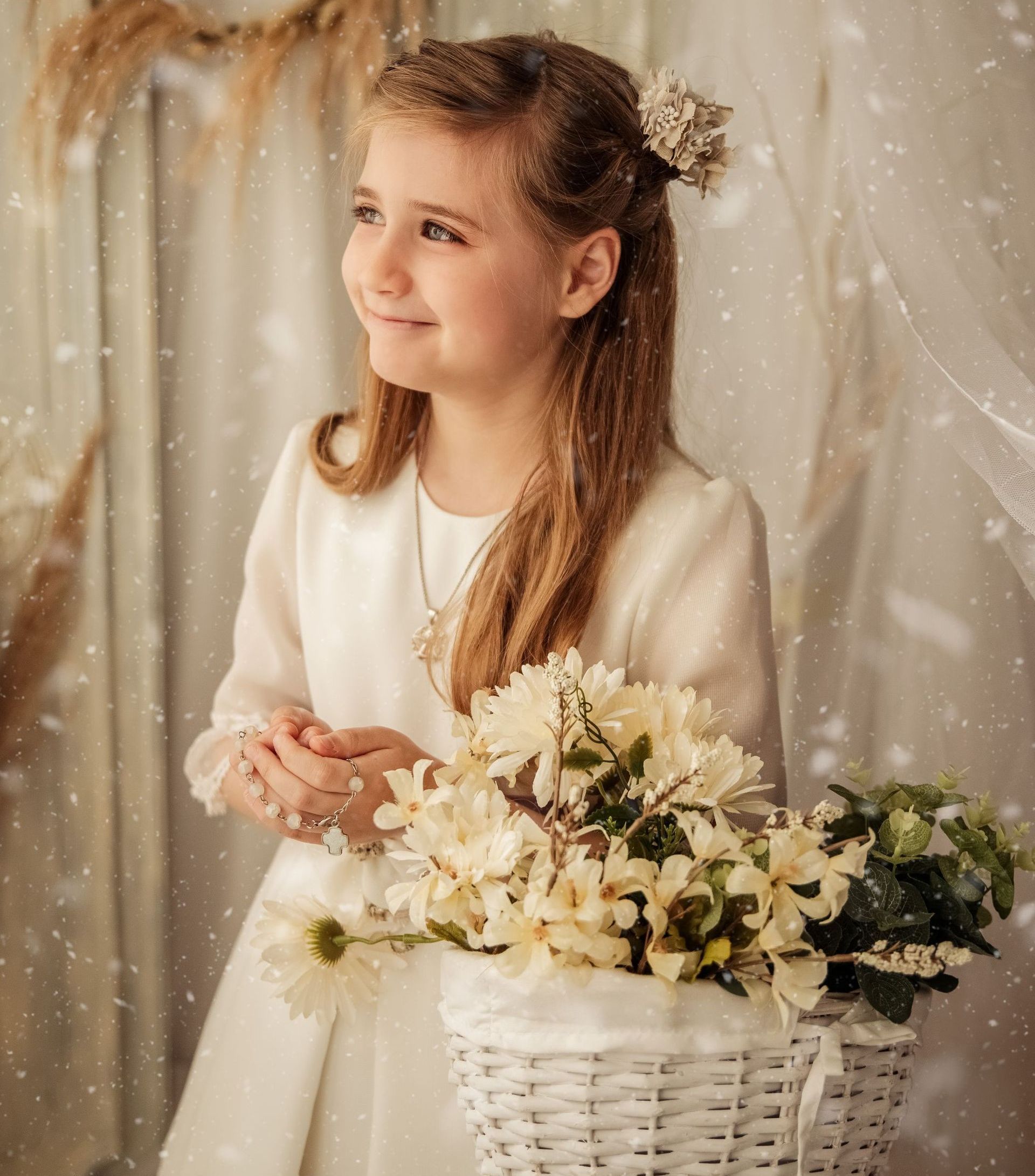 Niña con un vestido blanco sosteniendo flores junto a una cesta blanca de flores bajo un velo transparente.