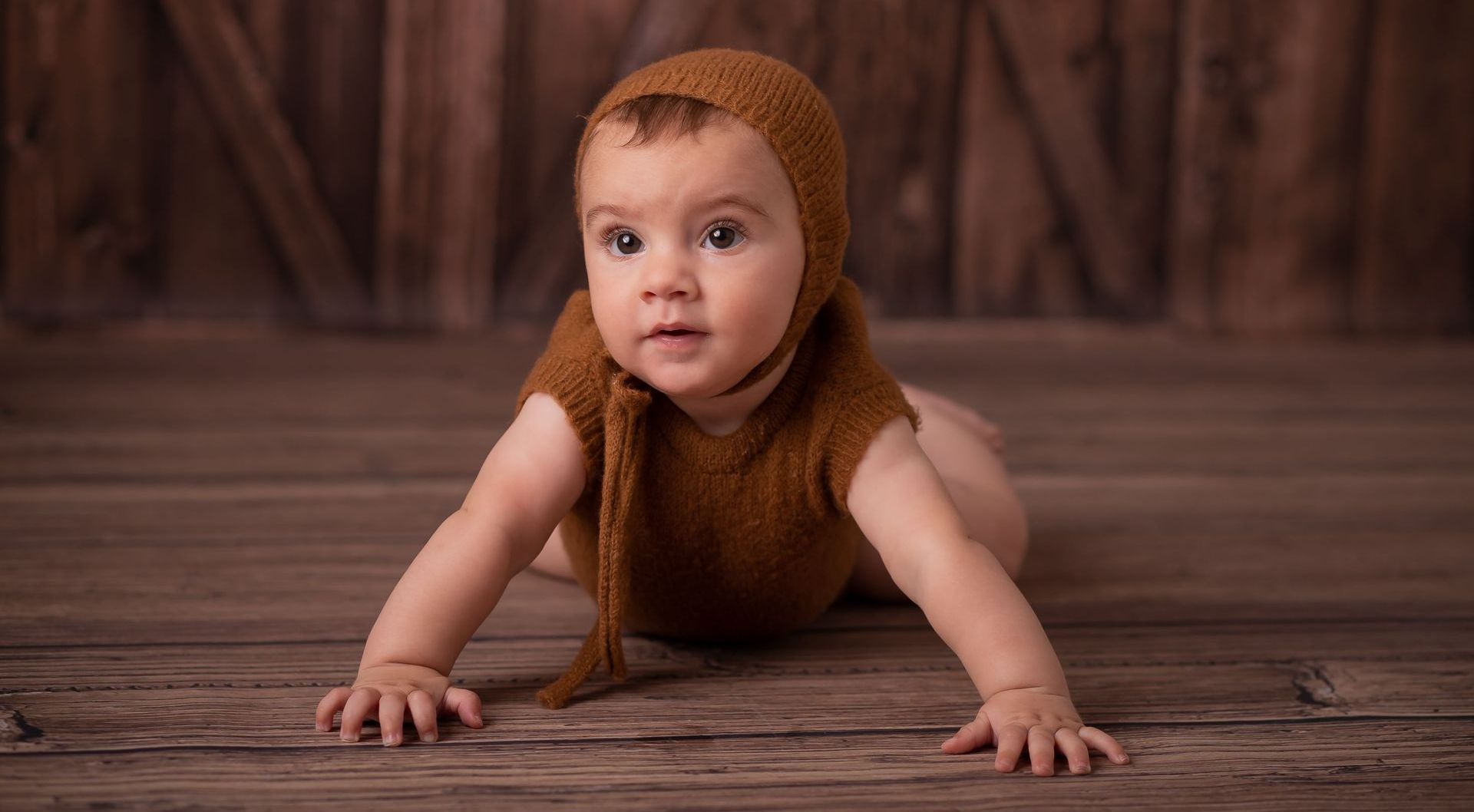 Un bebé con un gorro de punto marrón gatea sobre un suelo de madera, mirando hacia adelante.