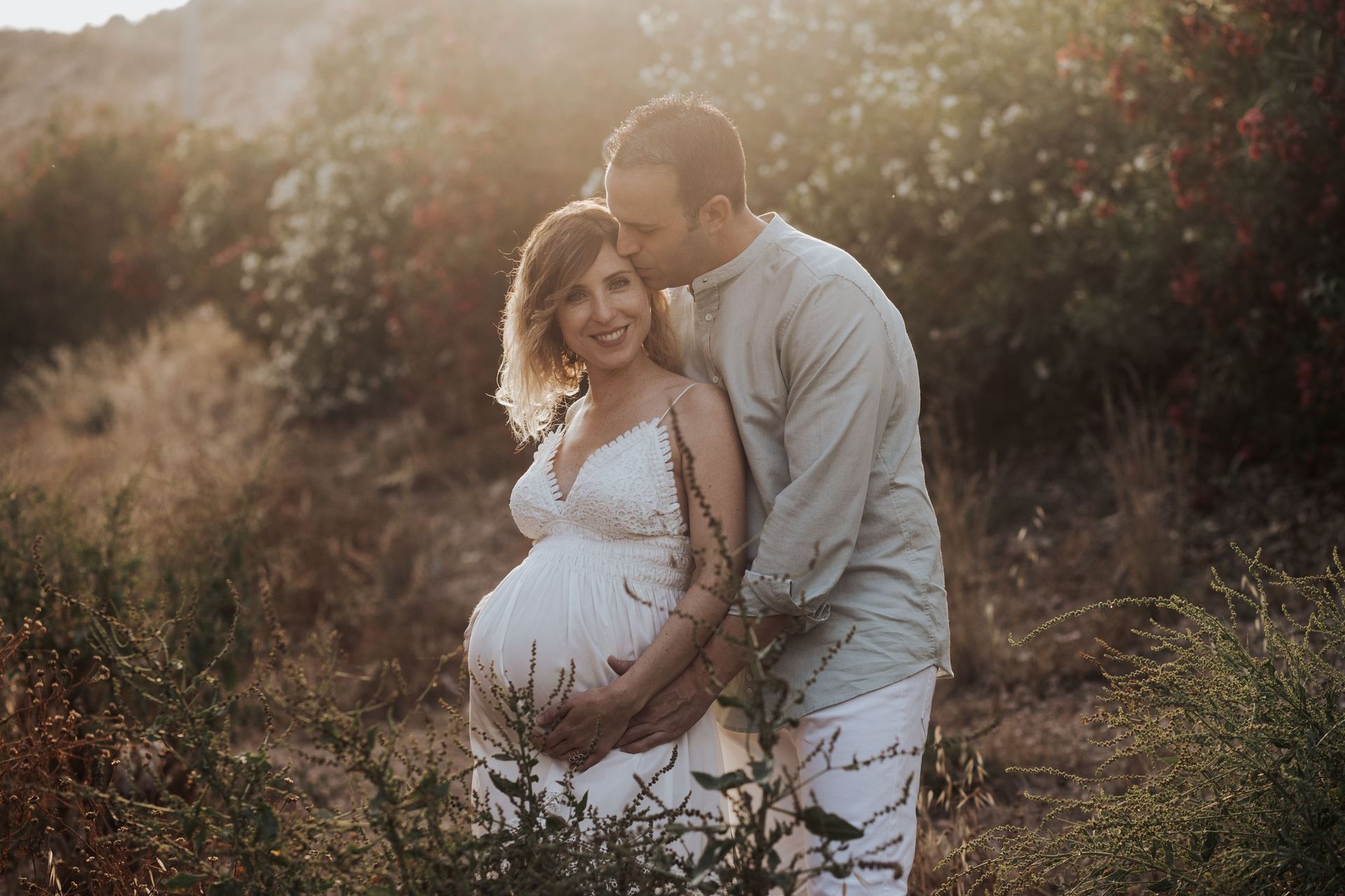 Una pareja embarazada se abraza al aire libre en un campo soleado, con una cálida luz dorada y una vegetación suave.