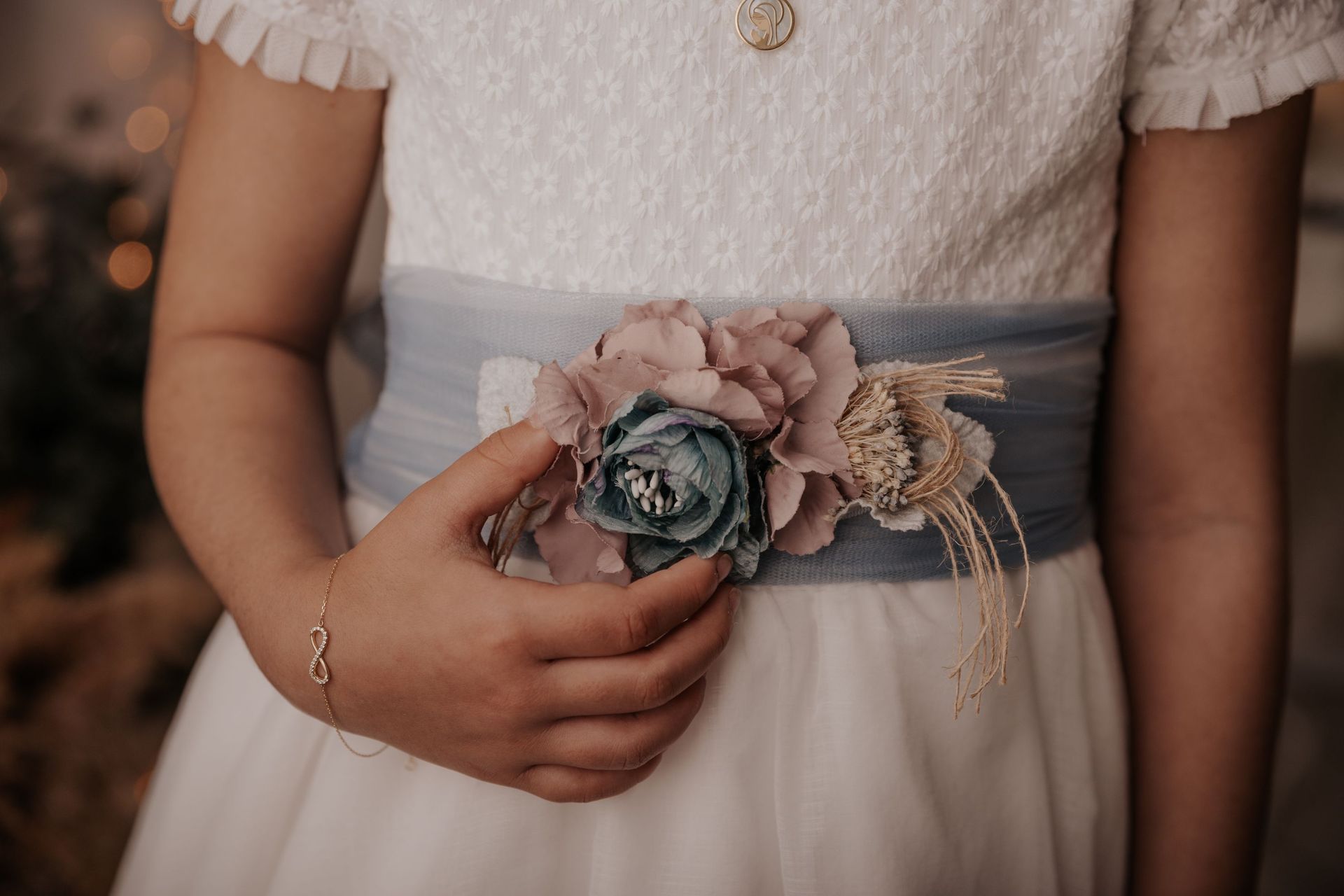 Manos de niña sosteniendo un ramillete floral sobre un vestido blanco con una faja azul.