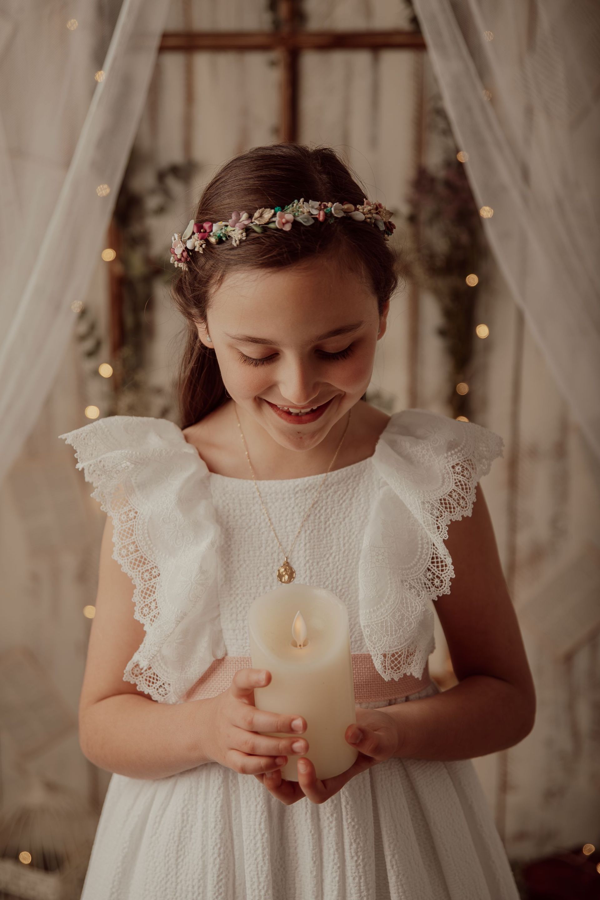 Una joven vestida de blanco sostiene una vela encendida y sonríe dulcemente en un ambiente cálido y festivo.
