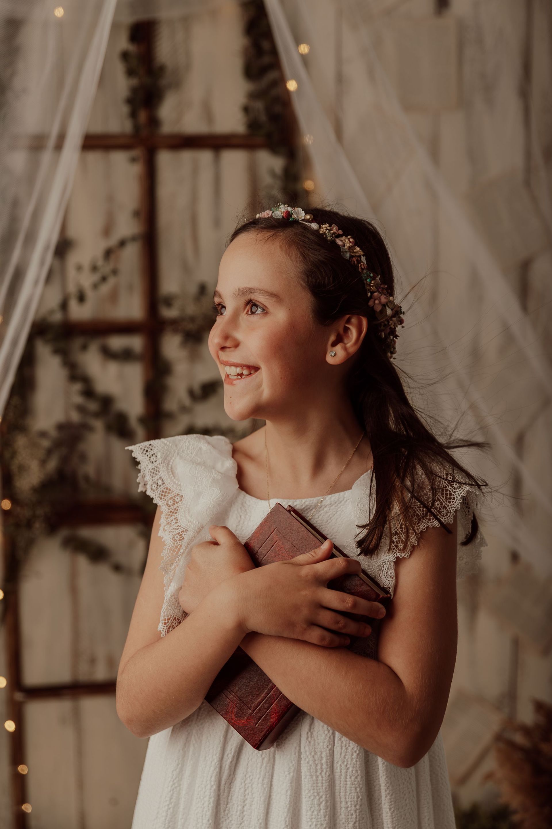 Niña sonriente con un vestido blanco, sosteniendo un libro y un regalo, de pie junto a una ventana con cortinas transparentes.