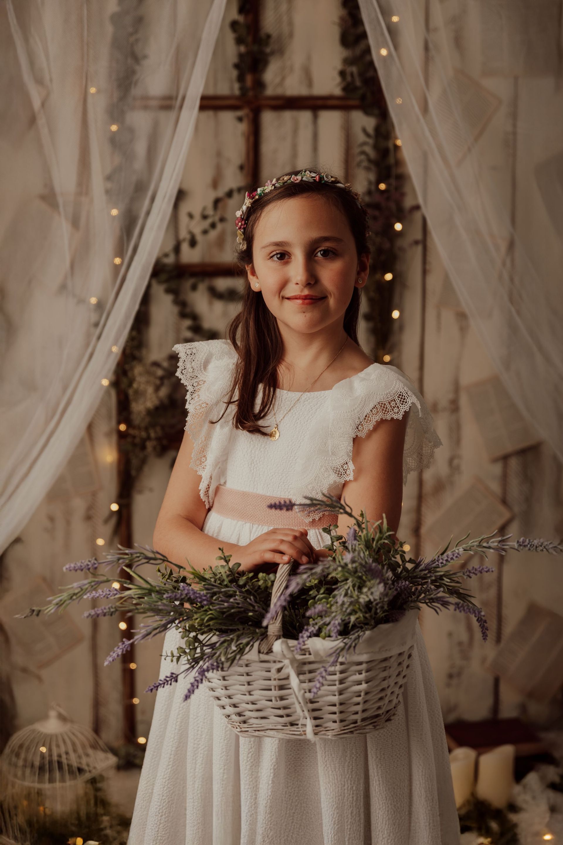 Una joven con un vestido blanco sostiene una cesta de flores, de pie frente a un fondo con una tela suave y drapeada.