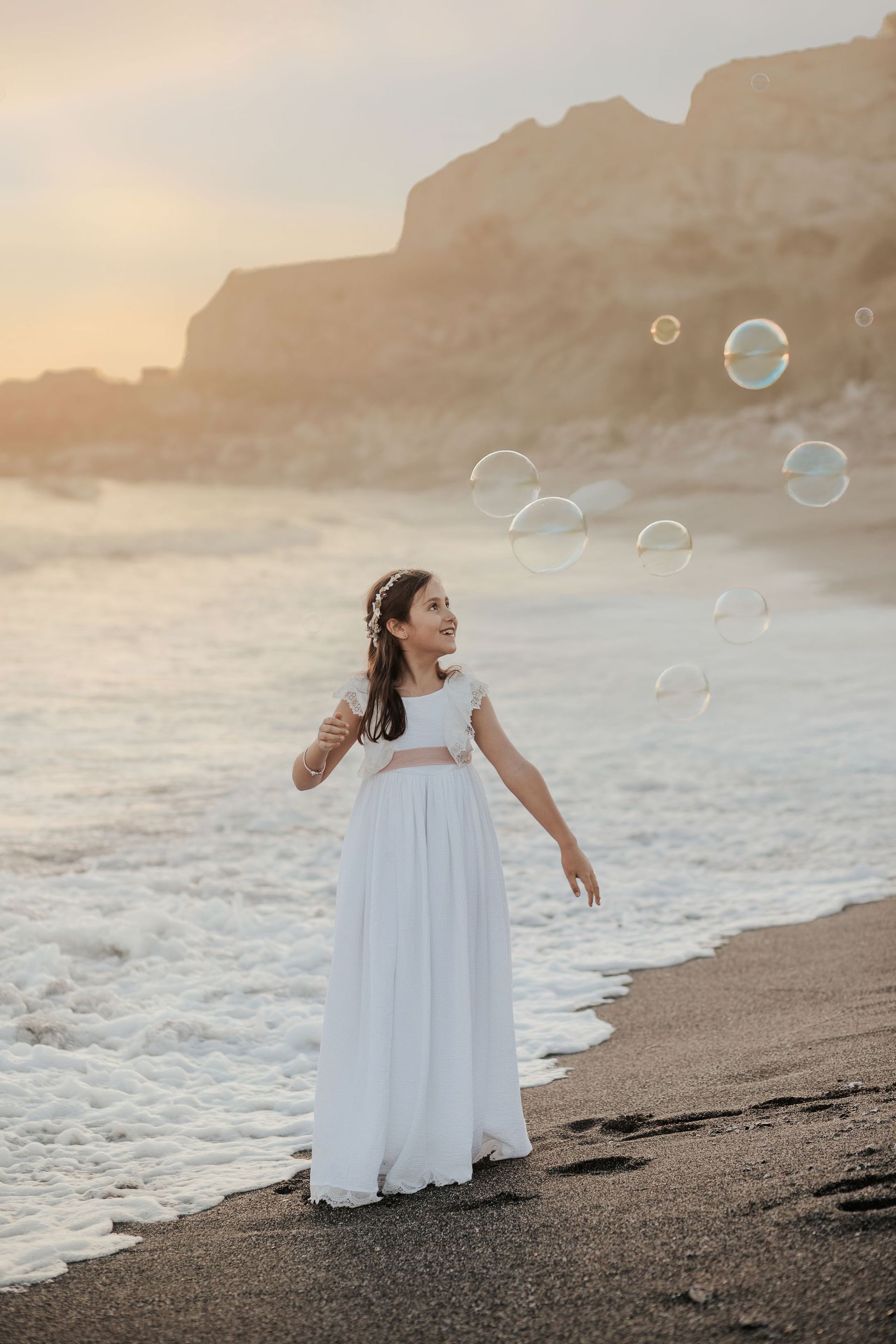 Mujer con vestido blanco de pie junto a la orilla al atardecer, soplando burbujas cerca de los acantilados marinos.