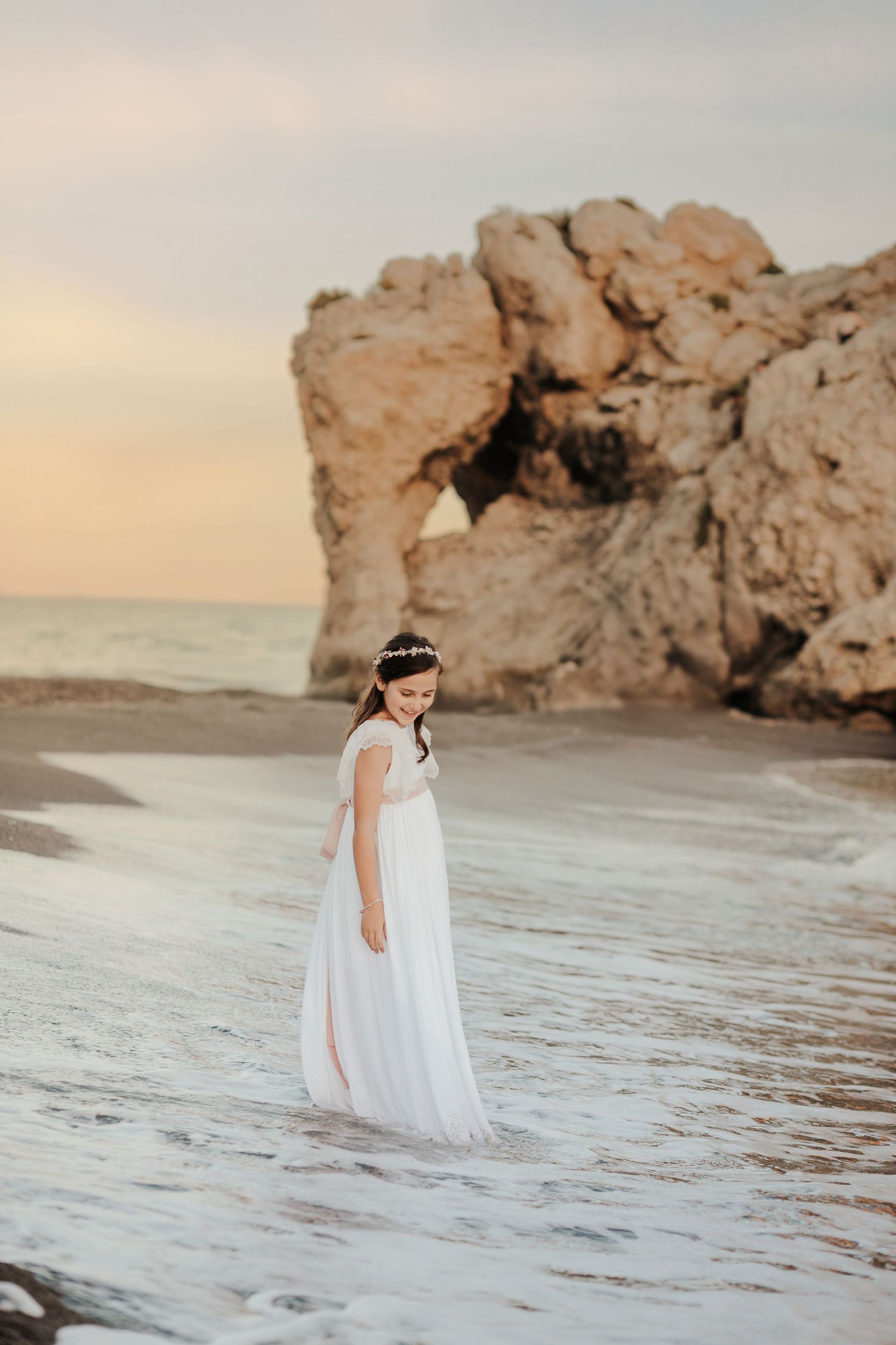 Mujer con vestido blanco de pie en una playa junto a un gran arco rocoso al atardecer.
