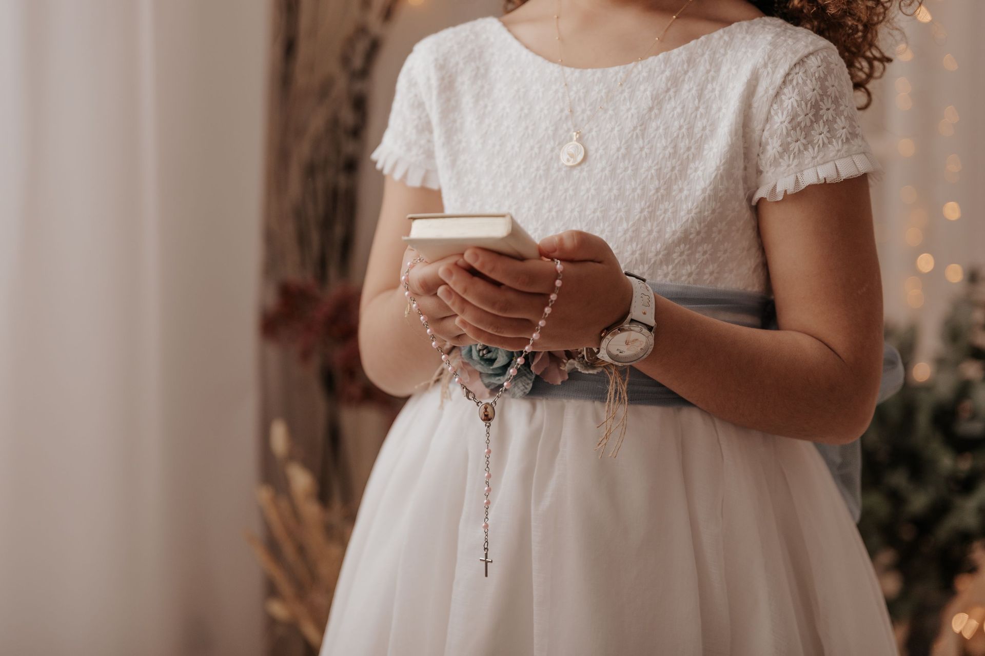 Mujer con vestido blanco sosteniendo un pequeño libro y un rosario en una habitación tenuemente iluminada.