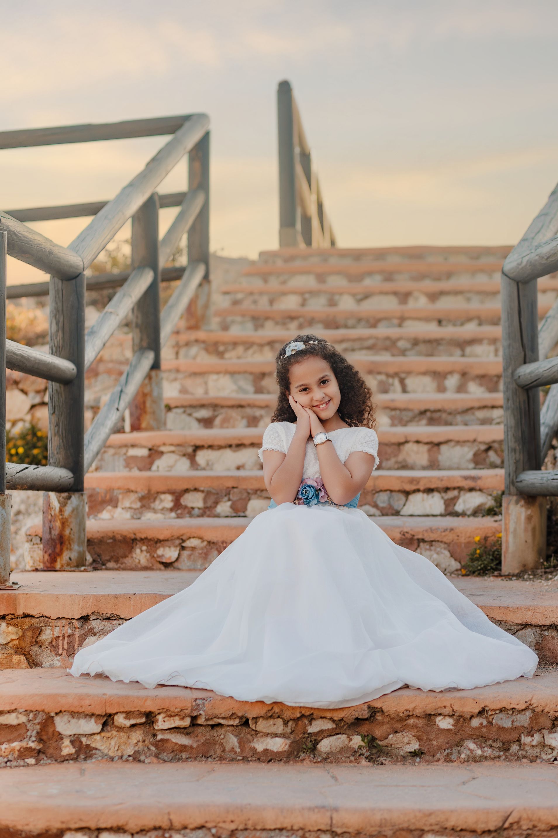 Una chica con un vestido blanco sentada en unas escaleras exteriores con barandilla al atardecer.