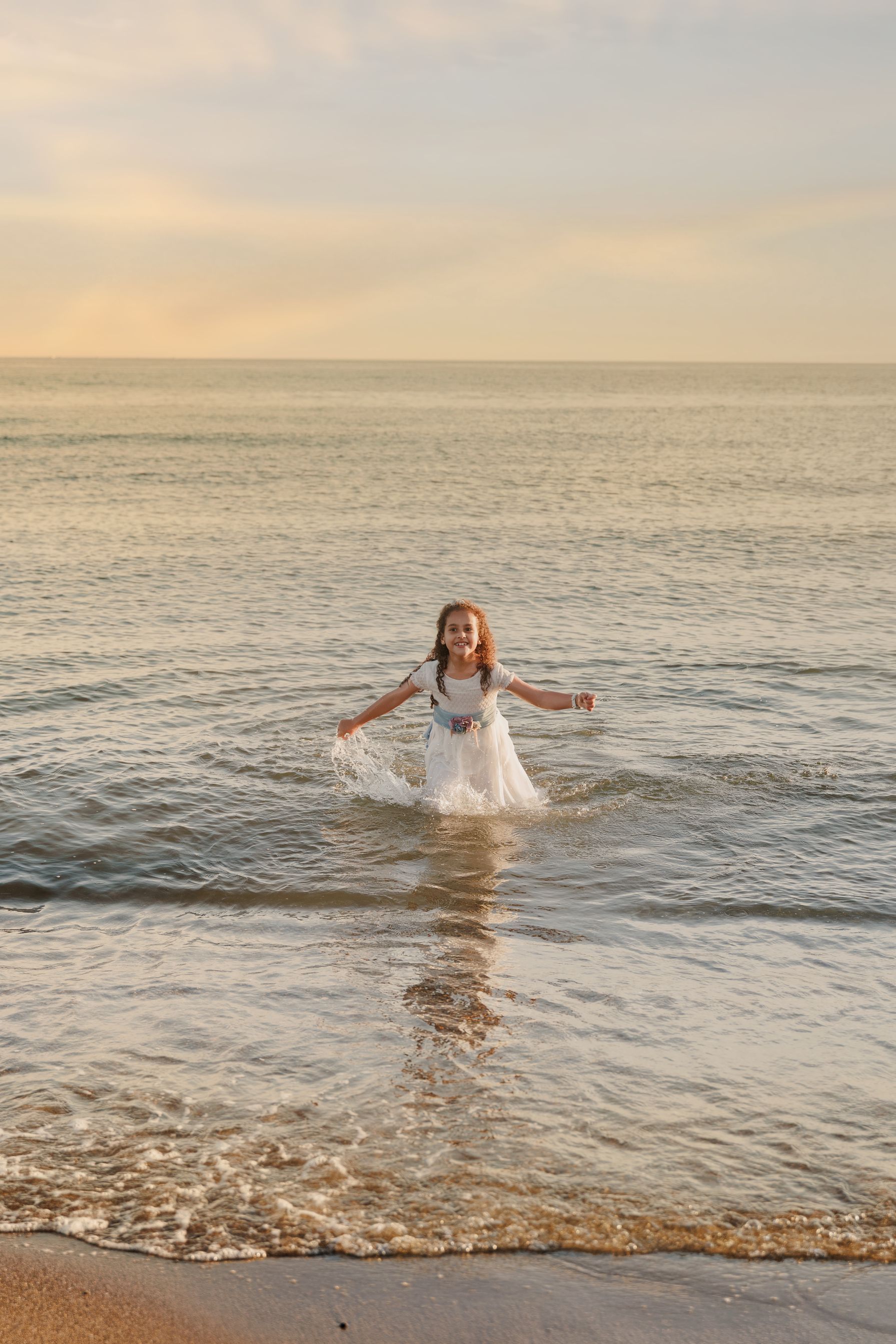 Persona vestida de blanco de pie en aguas poco profundas del océano al atardecer, con los brazos extendidos.