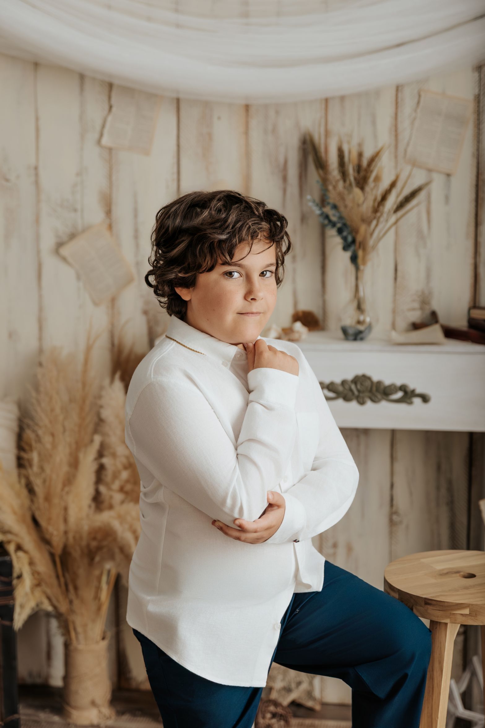 Persona sentada en un estudio de color beige, vestida con una camisa blanca y pantalones oscuros, posando con la mano en la barbilla.