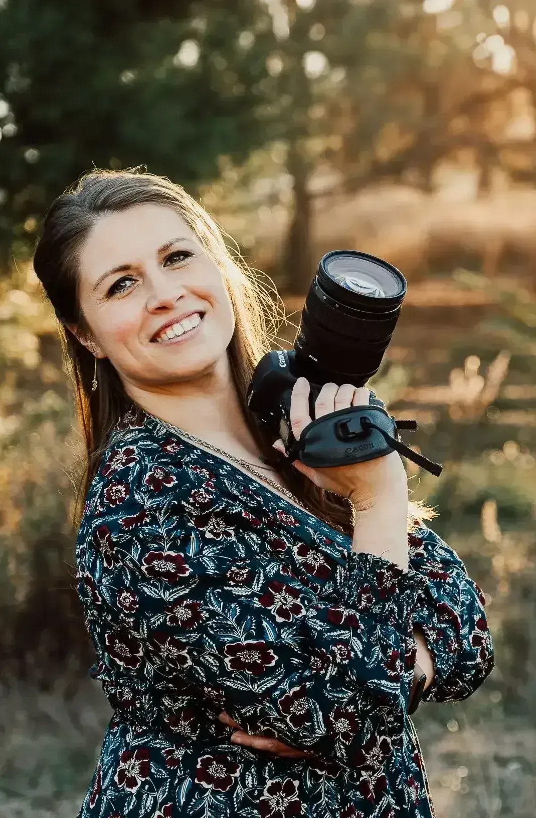 Mujer sonriente al aire libre, sosteniendo una cámara con un objetivo grande sobre su hombro bajo la cálida luz del sol.
