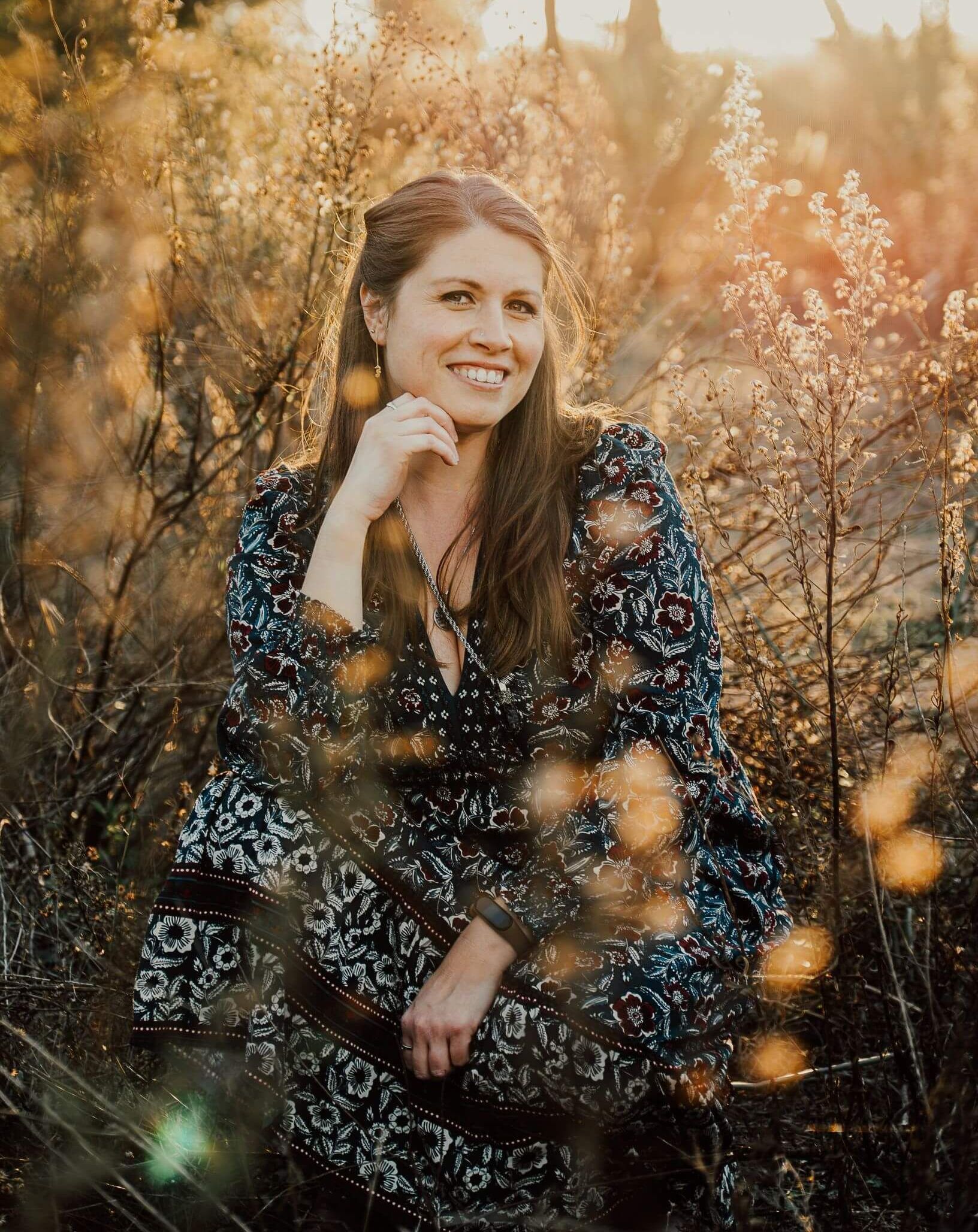 Mujer sonriente sentada en un campo soleado de hierba alta, vestida con un vestido oscuro estampado de flores.