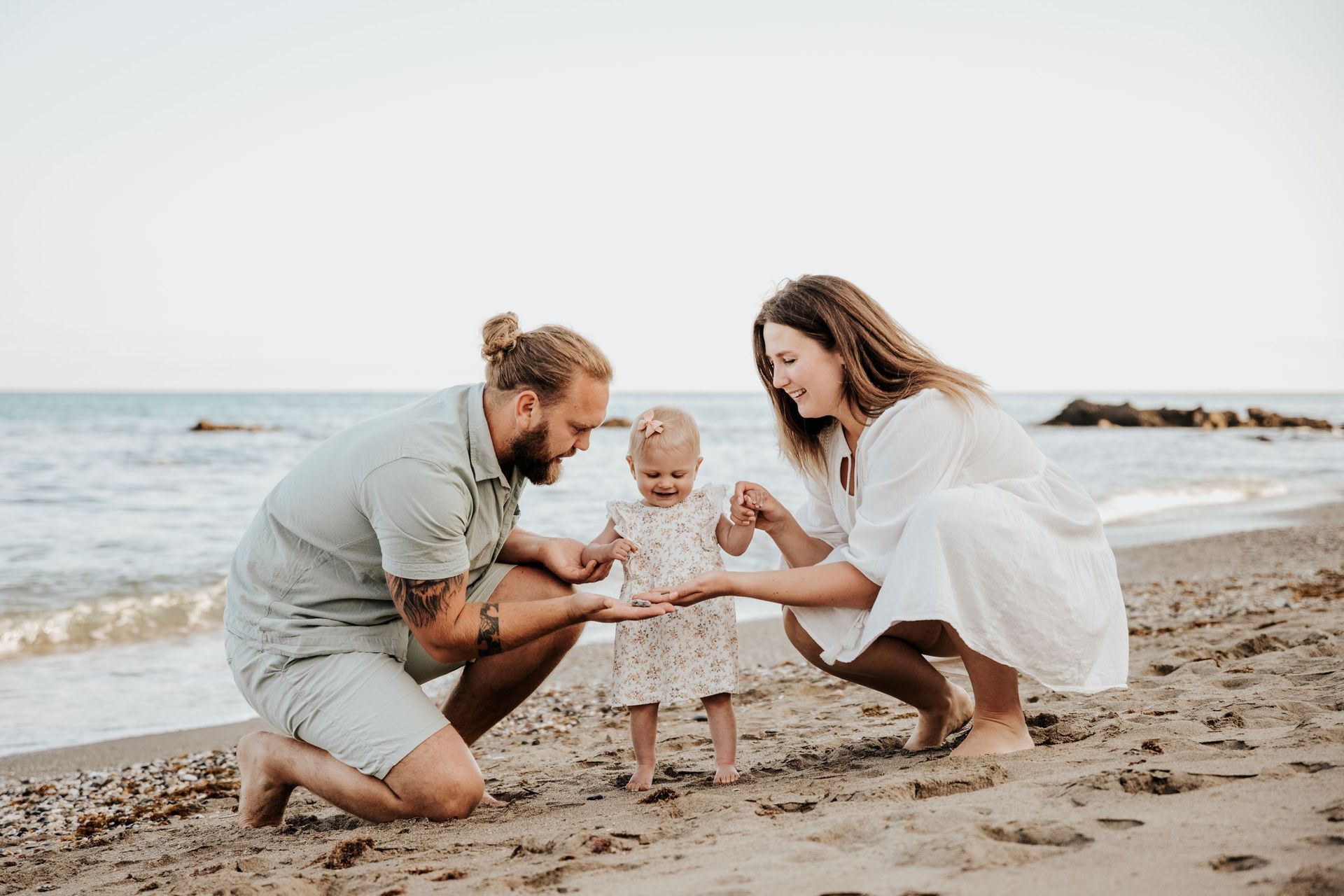 Una familia juega en la playa, a la orilla del mar, agachada alrededor de un niño pequeño junto al agua.