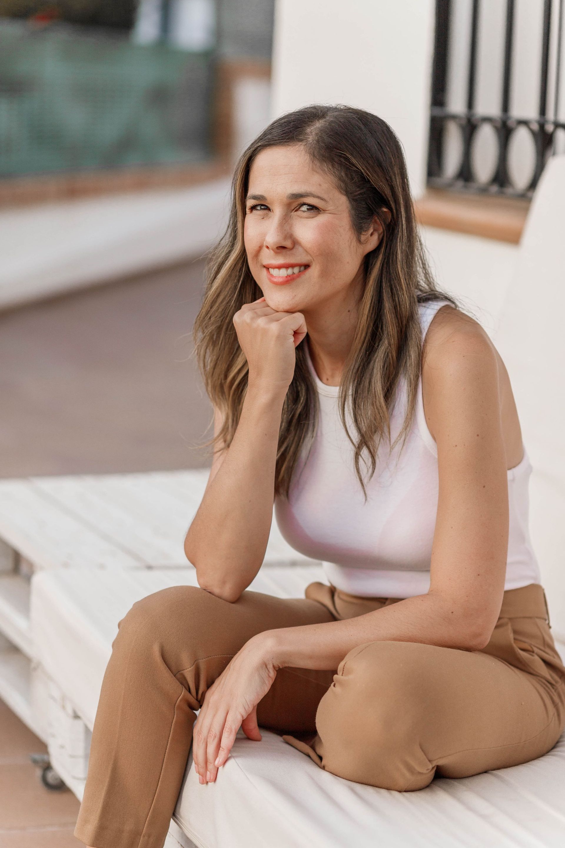 Mujer con camiseta blanca de tirantes y pantalones color canela sentada al aire libre, sonriendo con la barbilla apoyada en la mano.