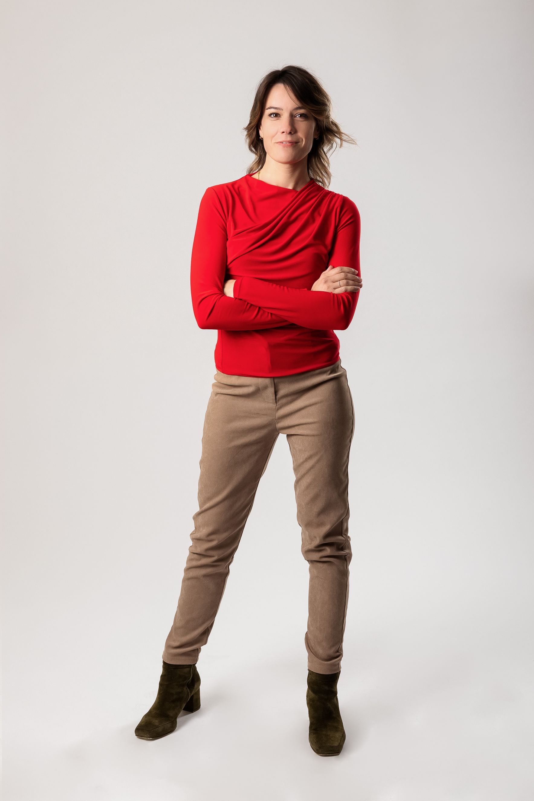 Persona de pie con los brazos cruzados, vestida con una blusa roja, pantalones color canela y botas oscuras sobre un fondo blanco liso.
