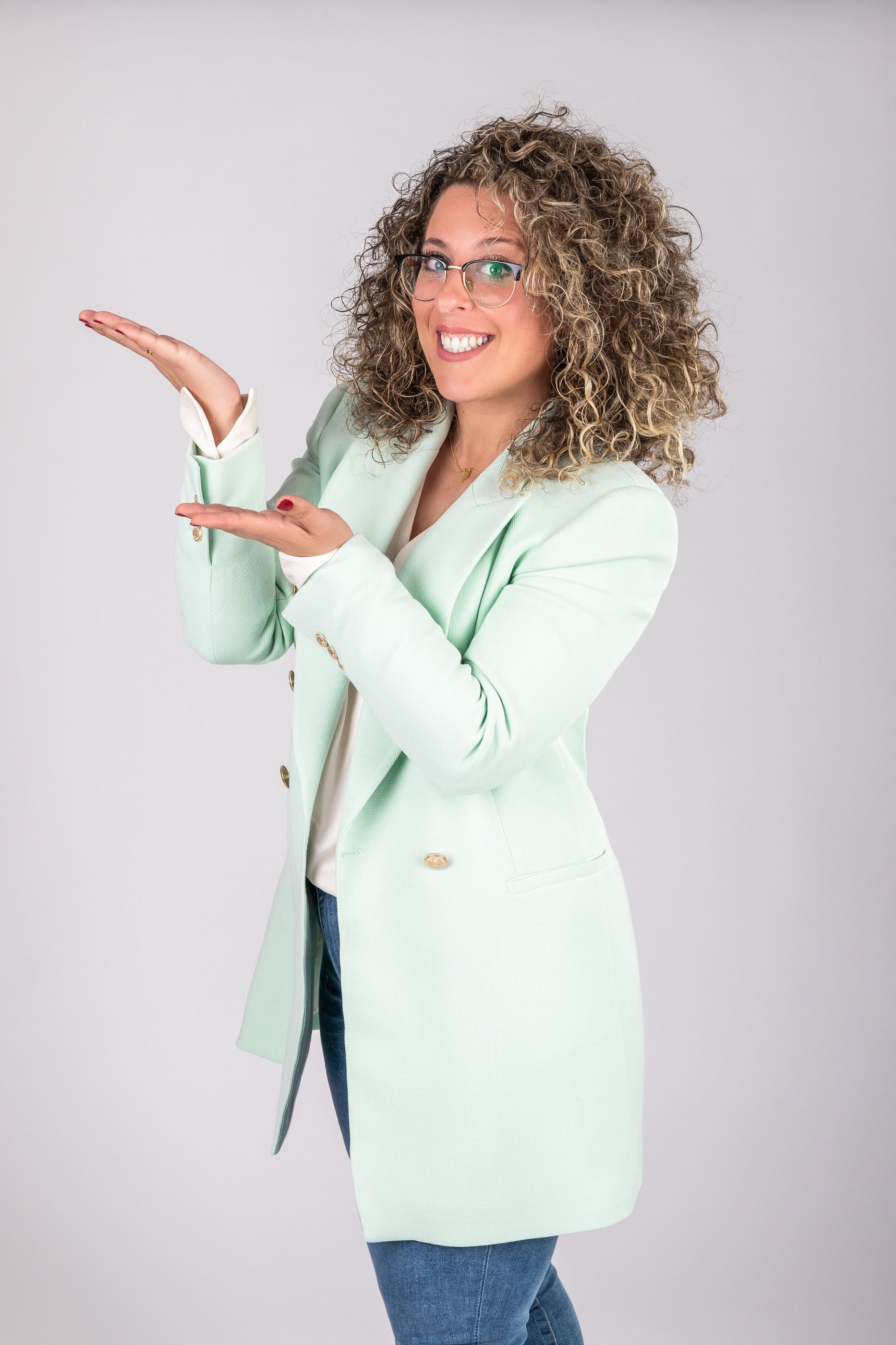 Mujer con blazer verde menta presentando con las manos abiertas sobre un fondo claro liso.