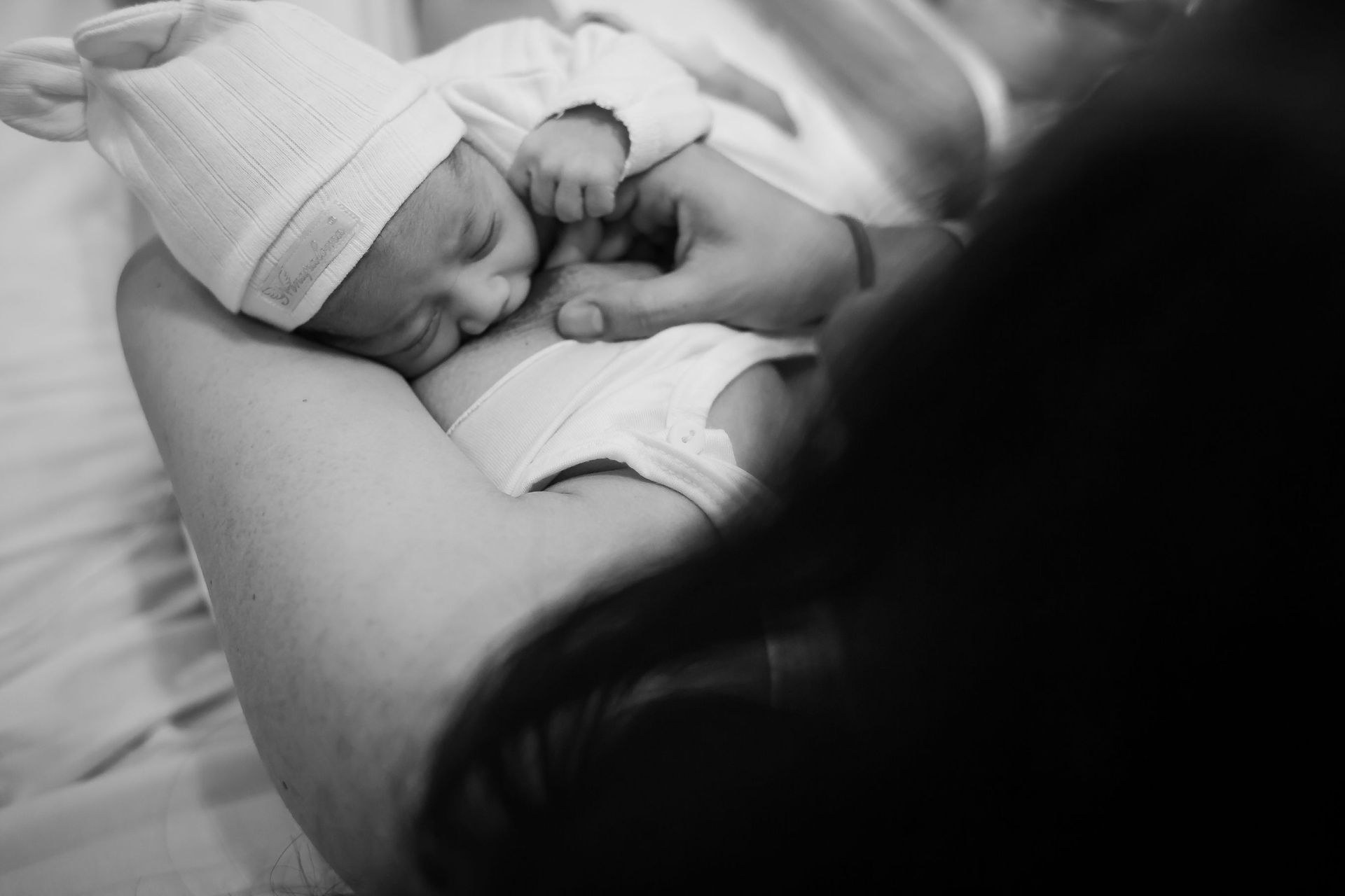 Un recién nacido durmiendo con un gorro de punto, acunado en brazos de un adulto en una suave escena en blanco y negro.
