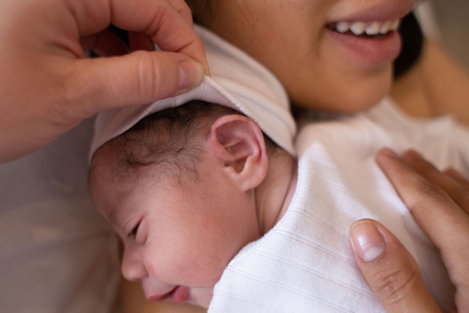 Un adulto sostiene con delicadeza a un bebé dormido, envuelto en una manta blanca, y le ajusta un gorro suave.