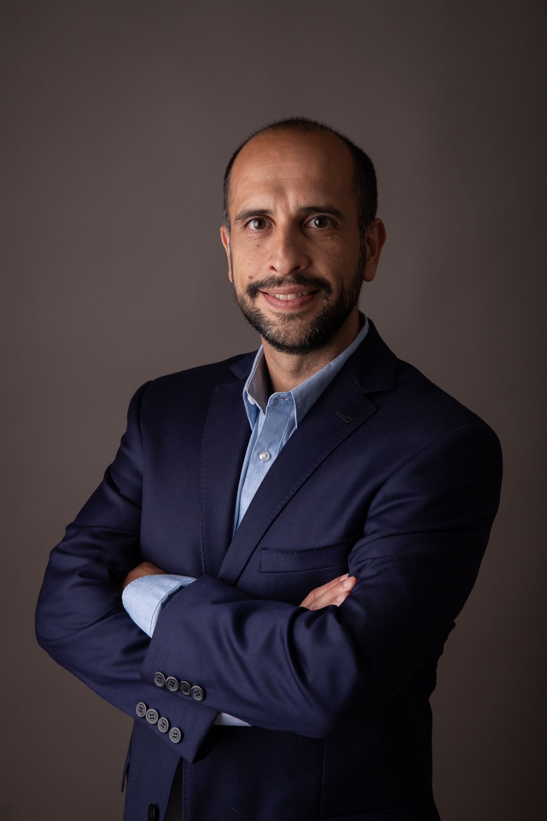 Retrato de un hombre con chaqueta azul marino y camisa azul, de pie con los brazos cruzados sobre un fondo oscuro.