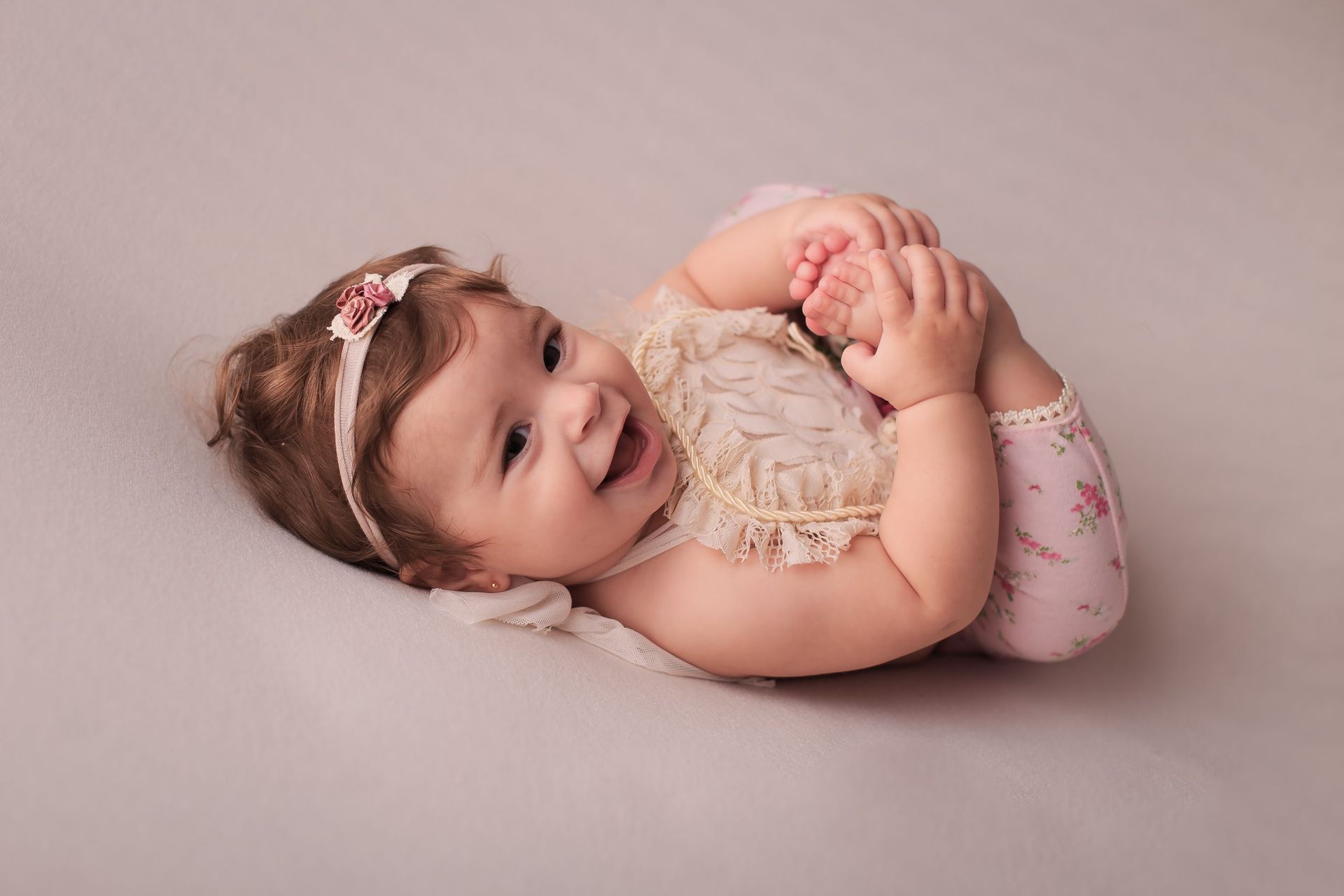 Un bebé sonriente yace sobre una manta rosa, sujetando sus pies y vistiendo un conjunto de color beige.