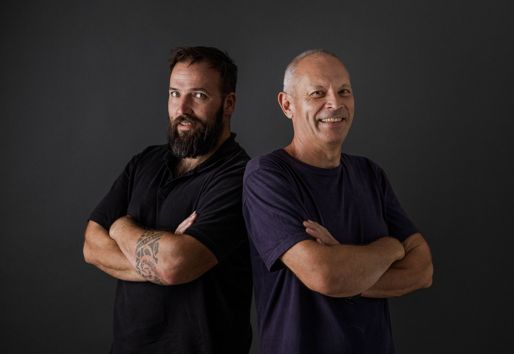 Dos hombres de pie espalda con espalda con los brazos cruzados sobre un fondo oscuro.