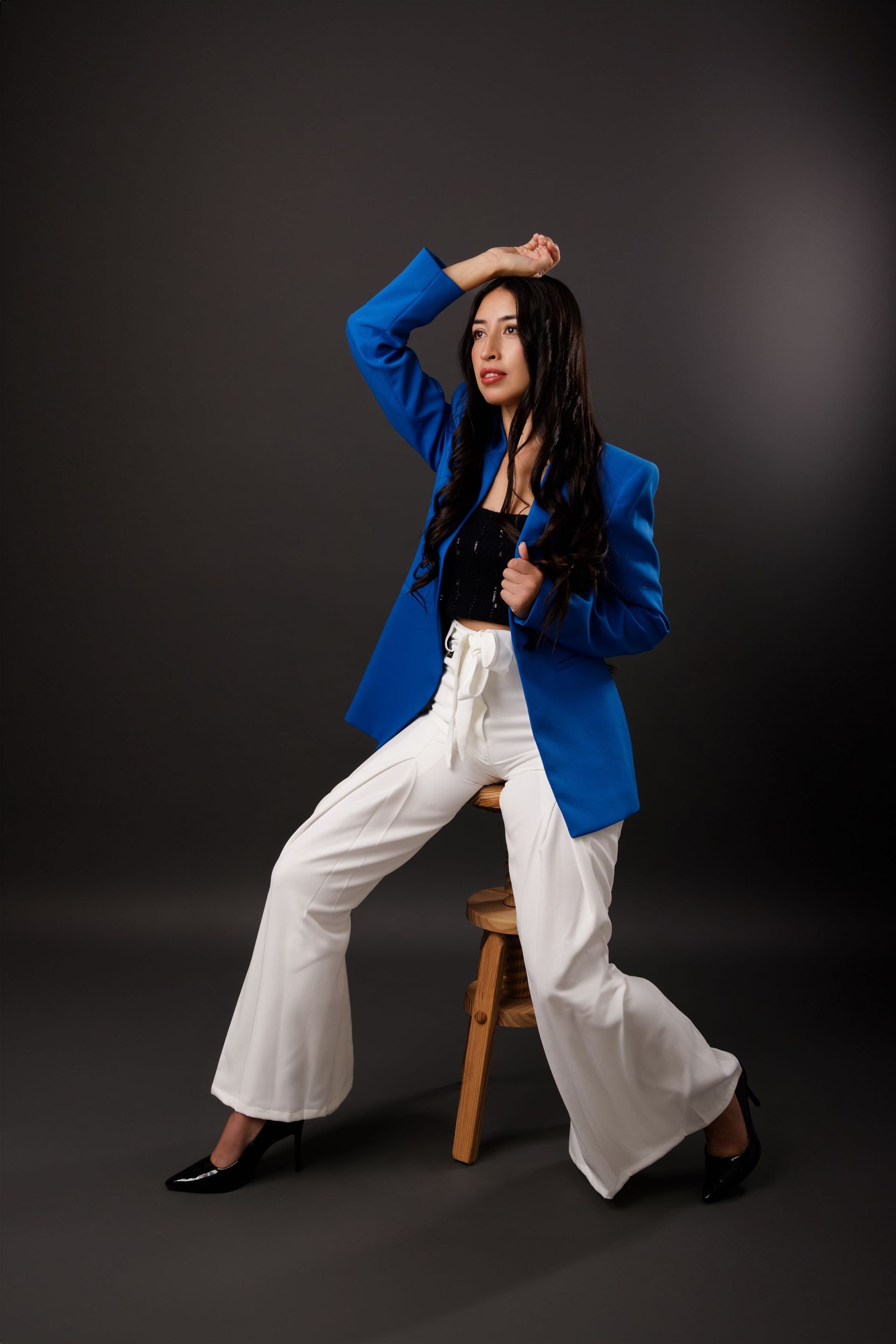 Mujer posando en un taburete con un blazer azul, pantalones blancos acampanados y tacones negros sobre un fondo oscuro de estudio.
