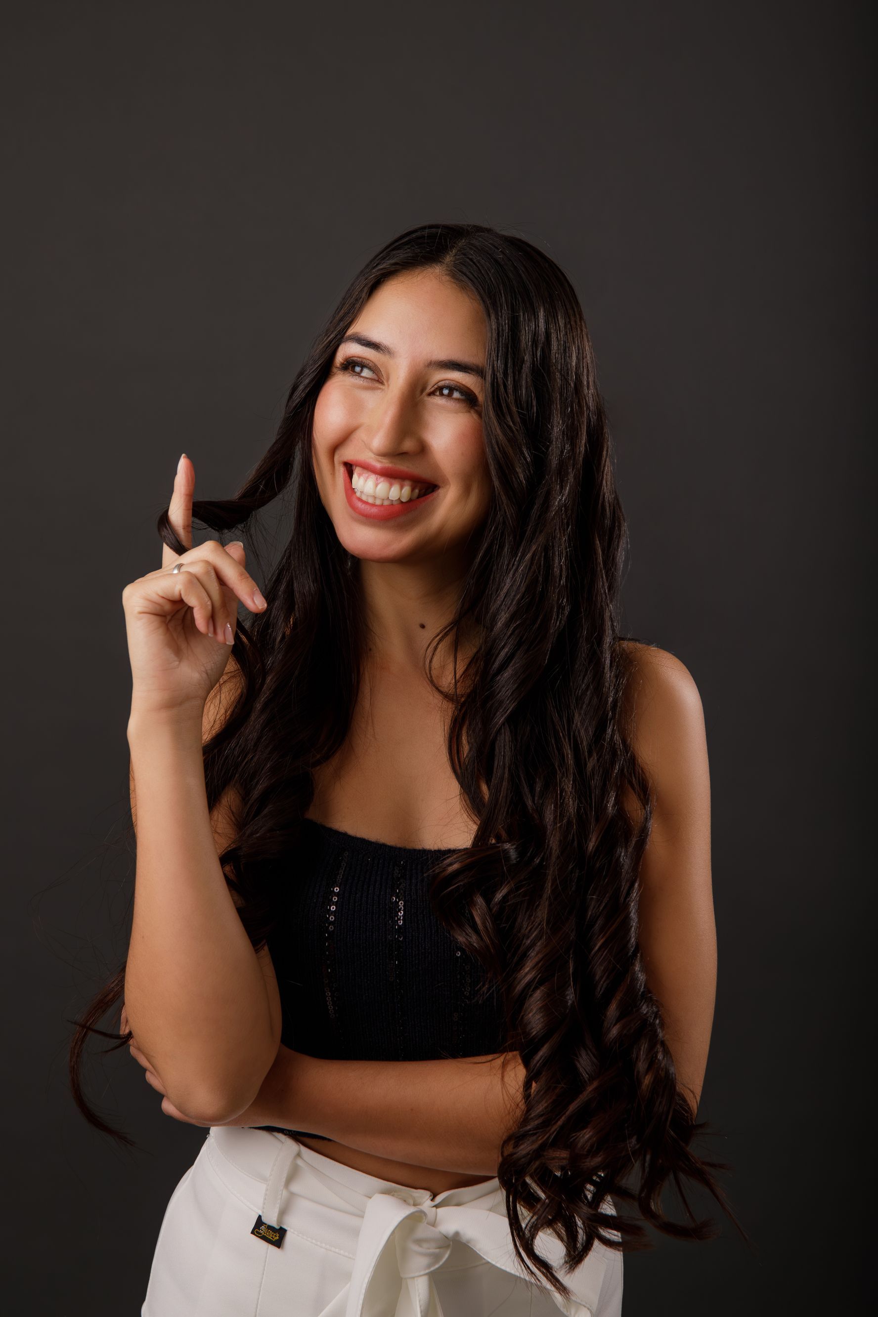 Una mujer sonriente, vestida con una blusa negra y pantalones blancos, señala hacia arriba sobre un fondo oscuro.