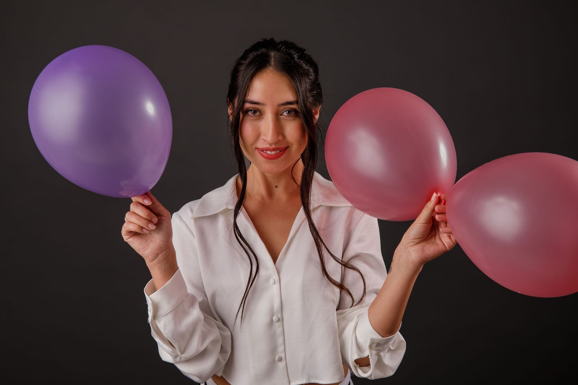 Mujer sosteniendo globos morados y rosas, sonriendo contra un fondo oscuro.