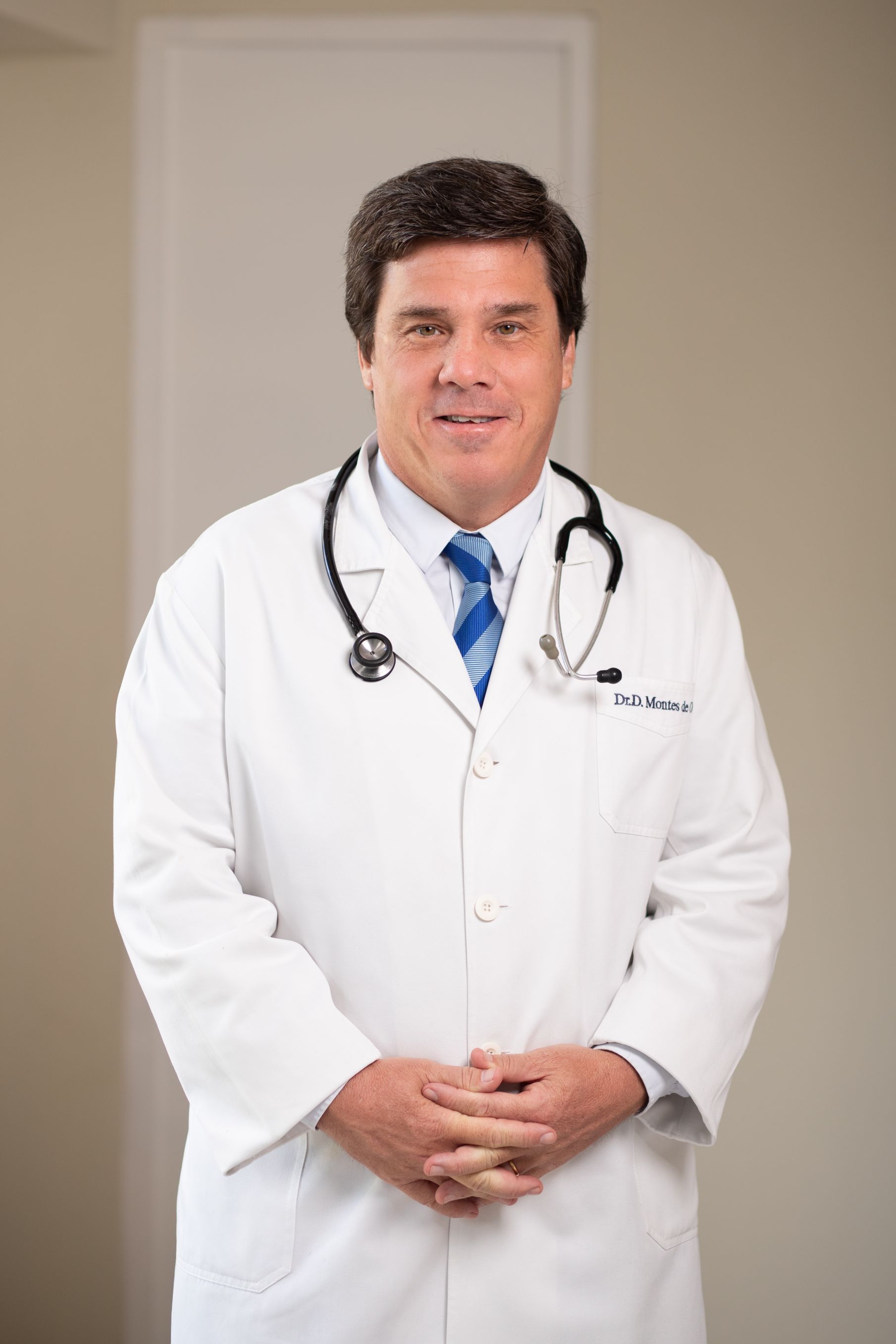 Un médico con bata blanca y estetoscopio, de pie en el interior de un edificio, con las manos entrelazadas.