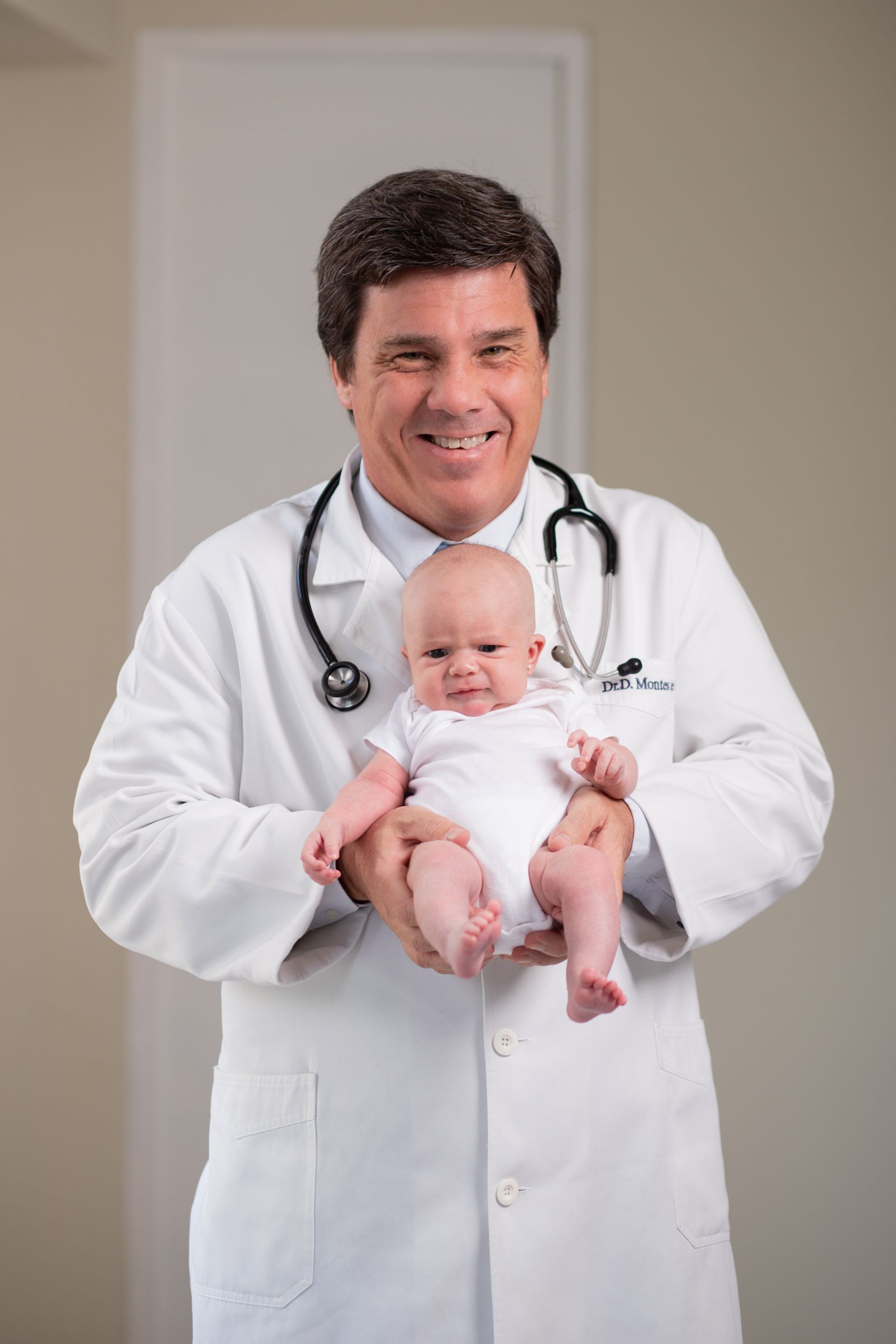 Médico con bata blanca sosteniendo a un bebé, sonriendo en un entorno clínico.