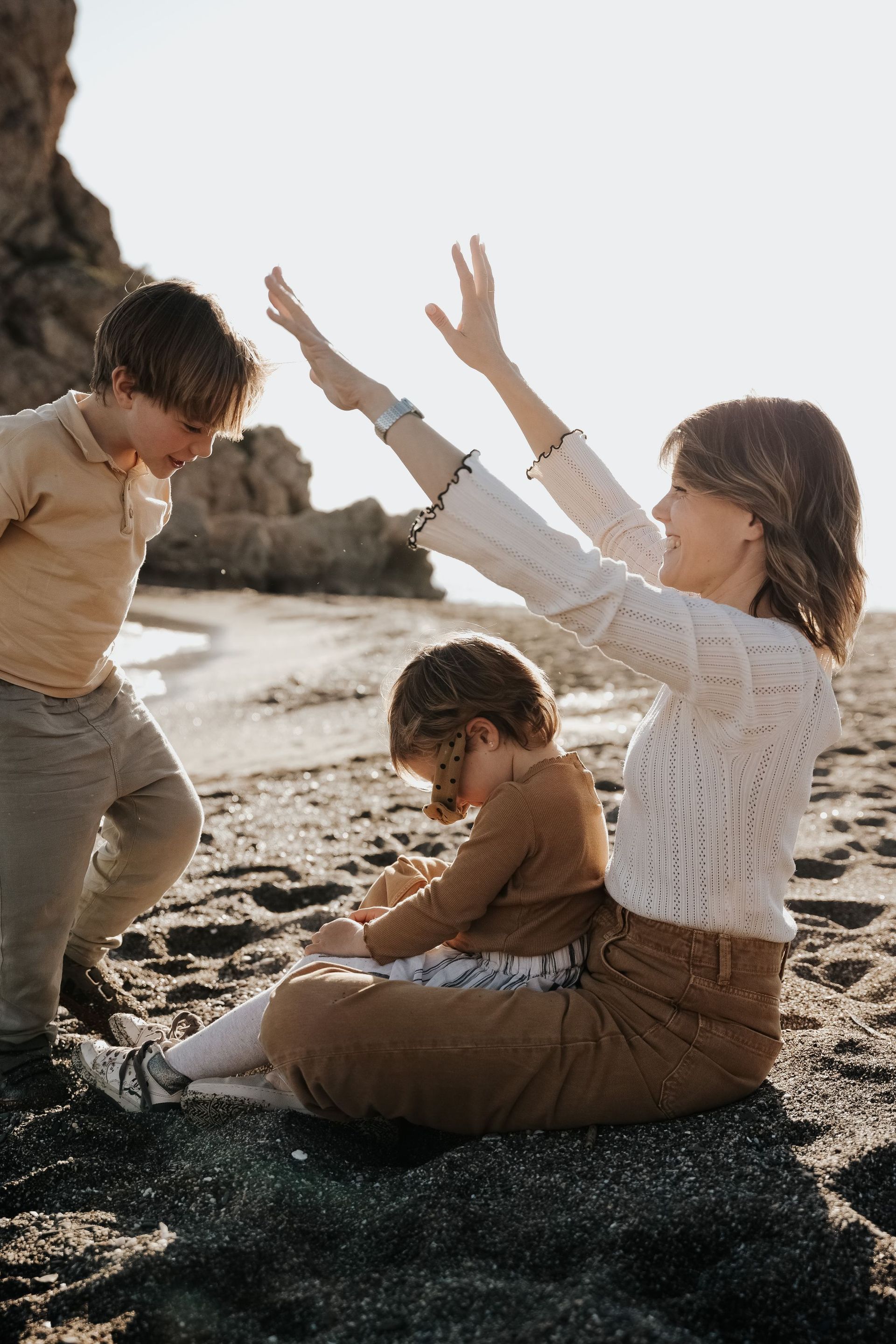 Dos niños en una playa, con un adulto levantando los brazos en un momento lúdico cerca del agua.
