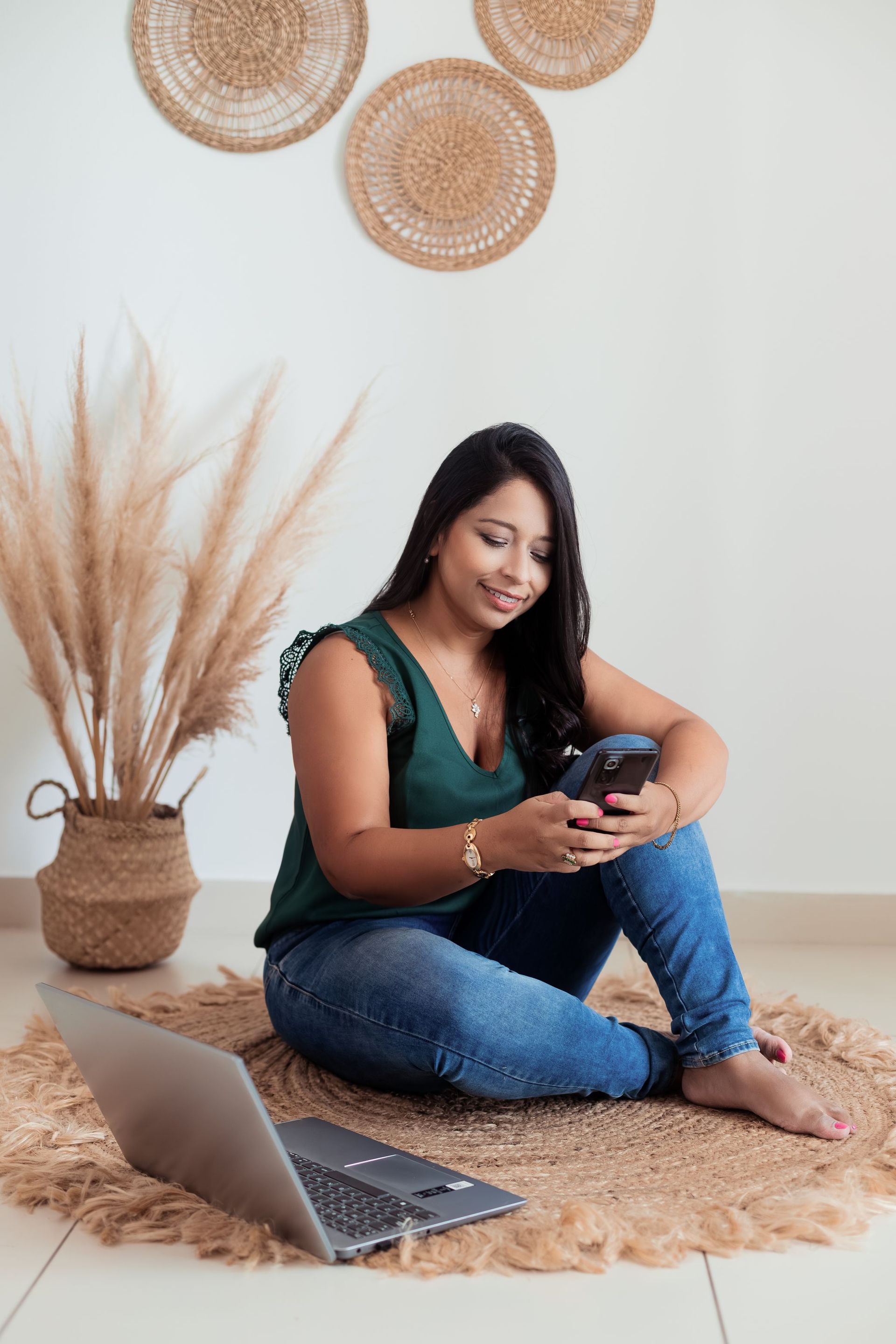 Una mujer sentada sobre una alfombra sonríe a su teléfono, que tiene al lado de un portátil abierto, en una acogedora habitación con decoración de tonos tierra.