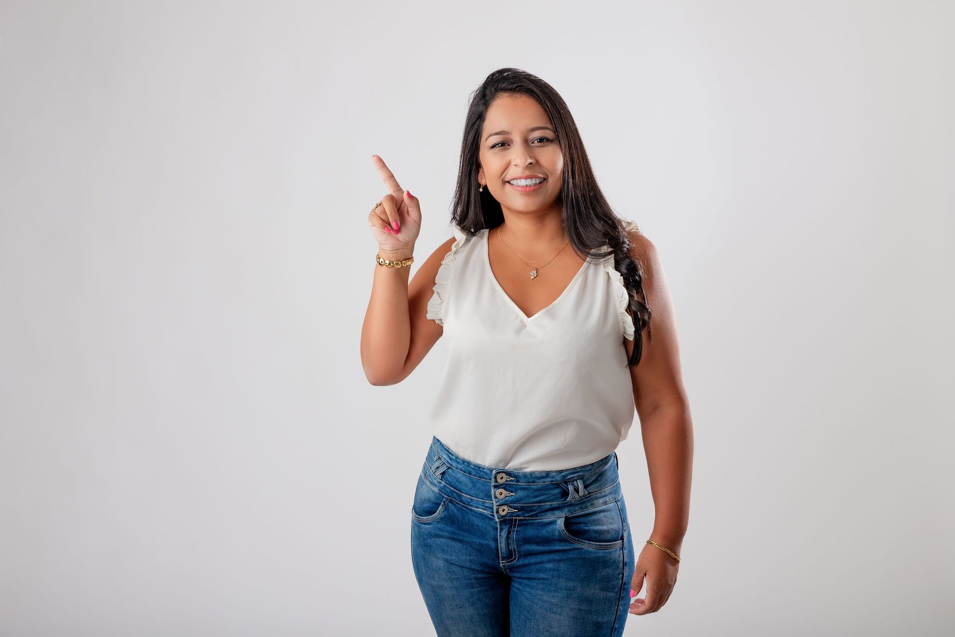 Mujer sonriente con blusa blanca y jeans señalando hacia arriba sobre un fondo gris liso.